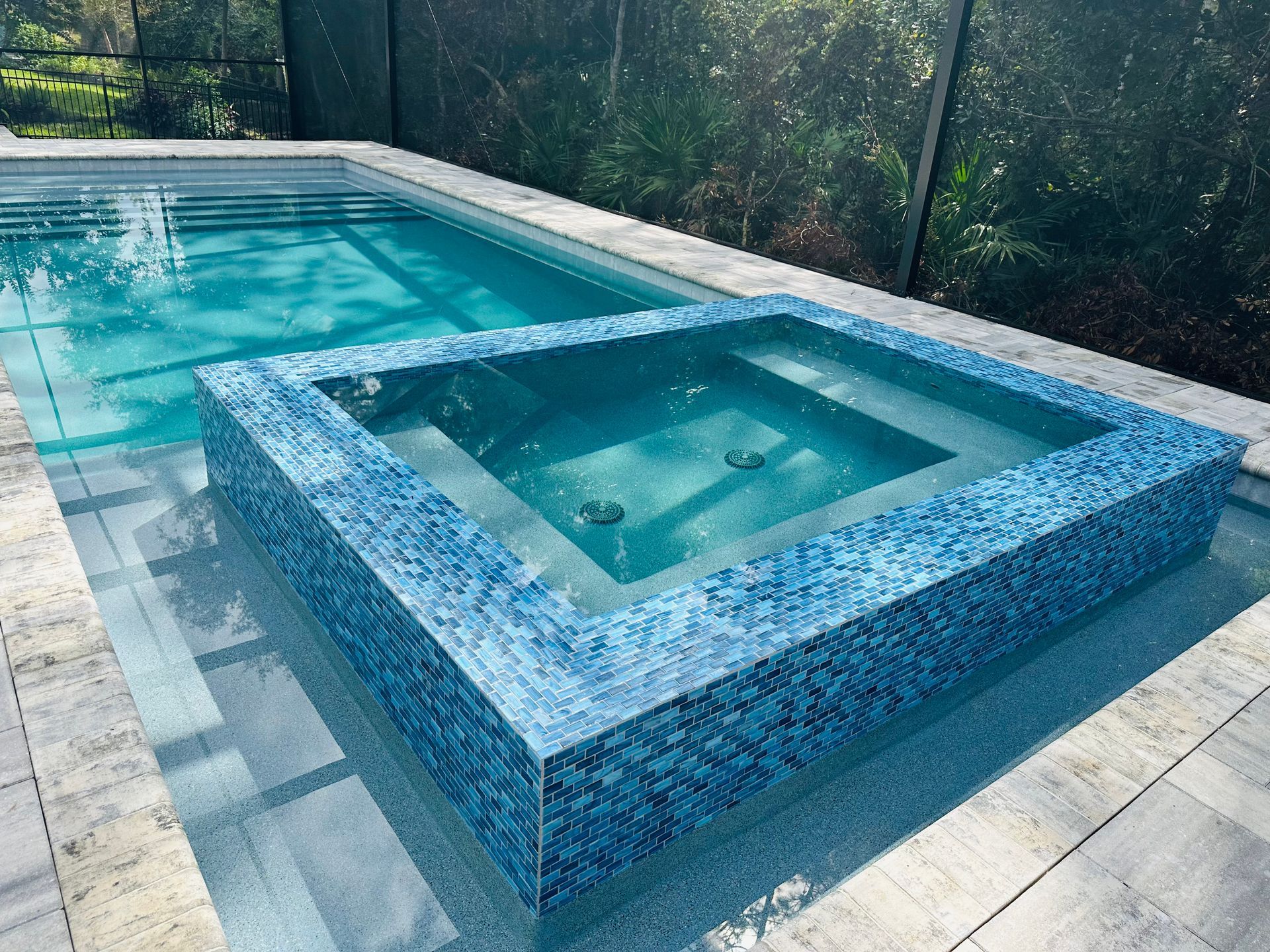 A modern swimming pool with a built-in spa, both featuring blue mosaic tile. Grey stone surrounds the pool.