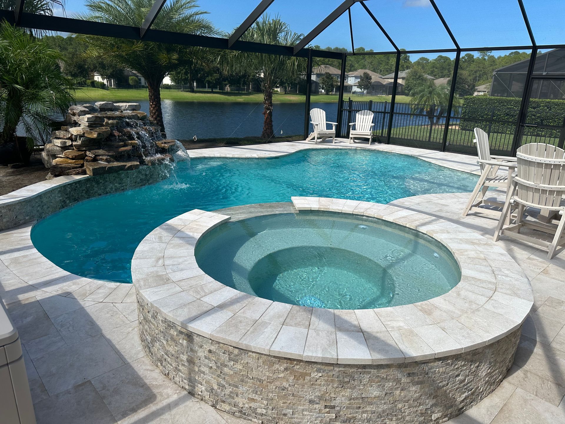 A swimming pool with a waterfall and spa on a patio.  Blue water, light-colored stone, and views of a lake and trees.
