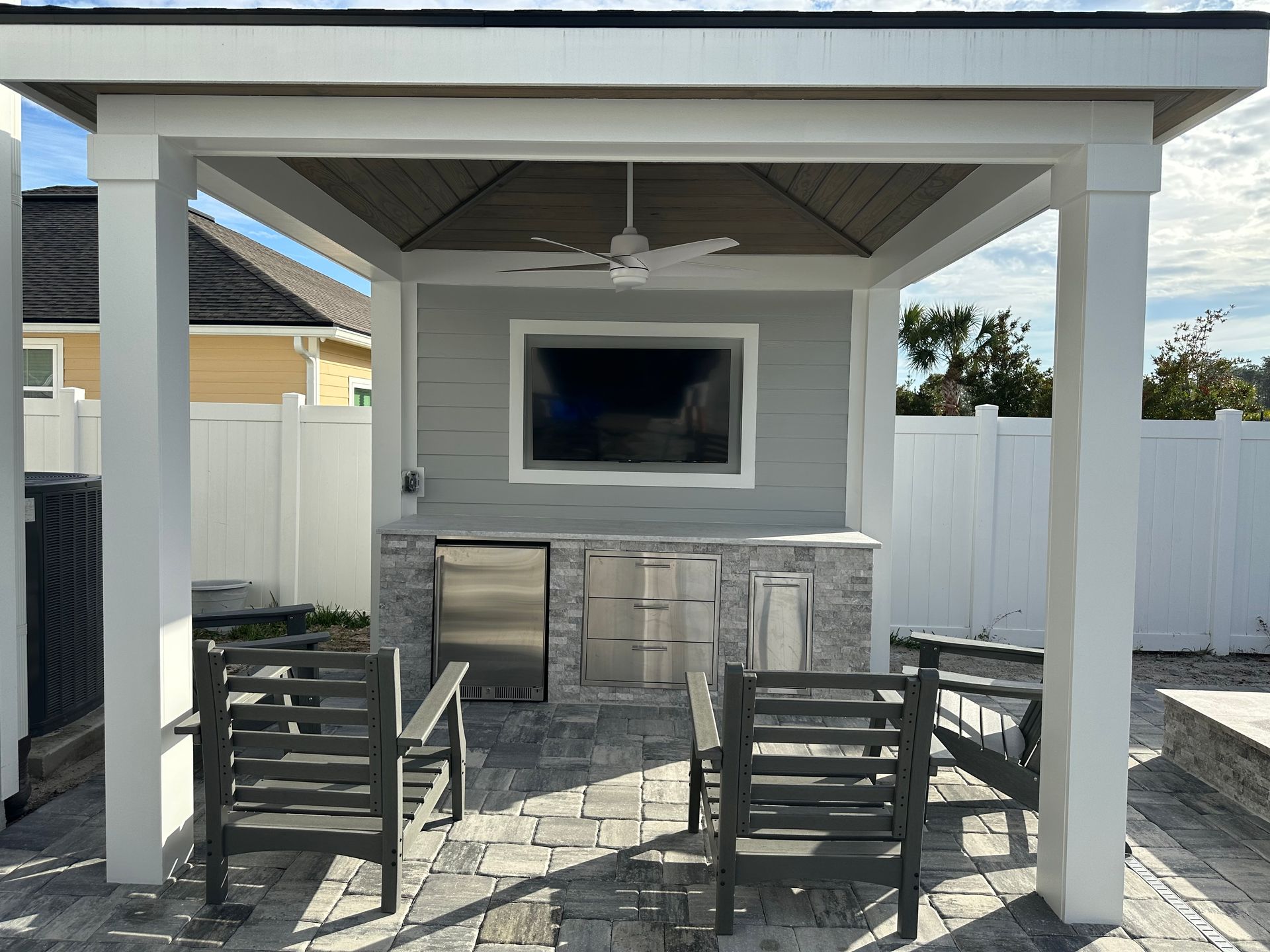 Outdoor kitchen with seating. Features a gray built-in structure with a TV, grill, and fridge, under a white-framed roof.