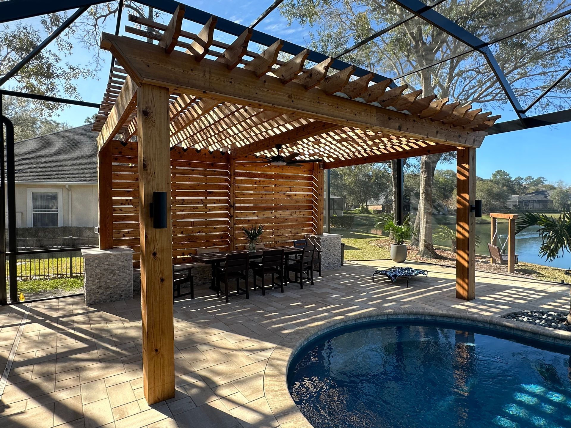 Pergola with slatted wood walls and a dining table, beside a swimming pool overlooking a lake.