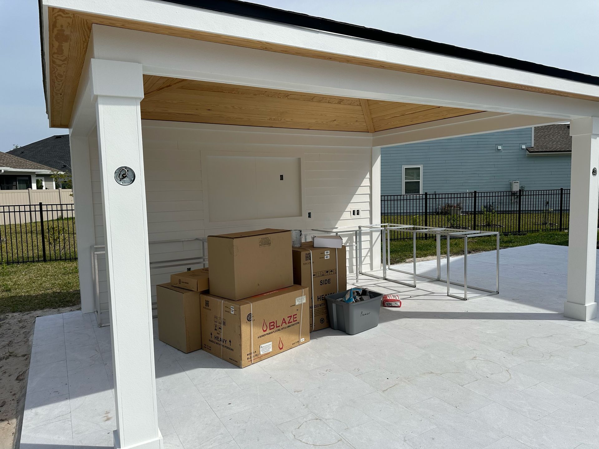 Boxes and equipment sit under a white-painted outdoor structure with a light-colored ceiling, next to a patio with grass and a fence.