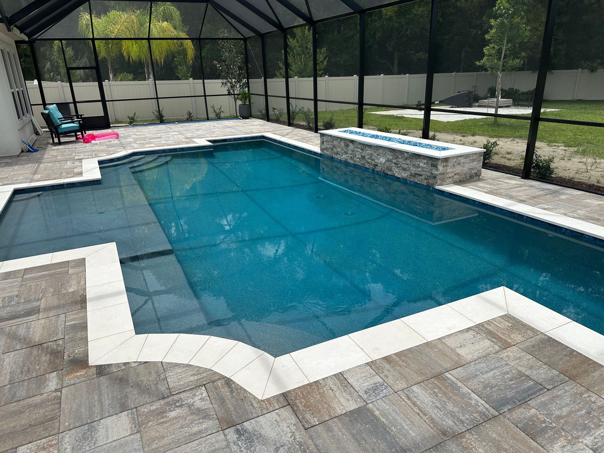 A rectangular swimming pool with a fire pit, surrounded by grey stone pavers and a screened enclosure.