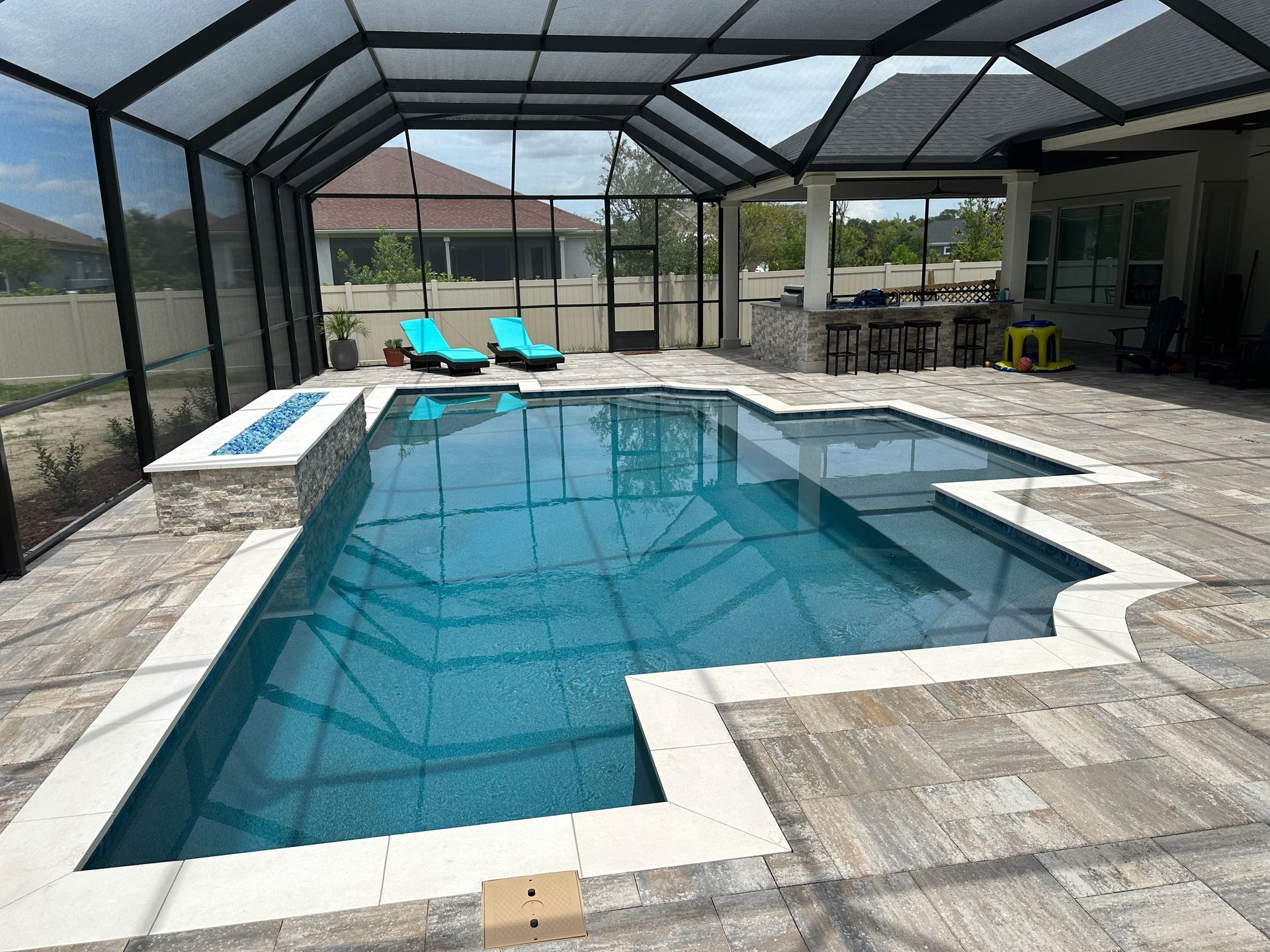 A pool enclosed by a screen. Two blue lounge chairs sit by the pool. Paved patio with a bar area.