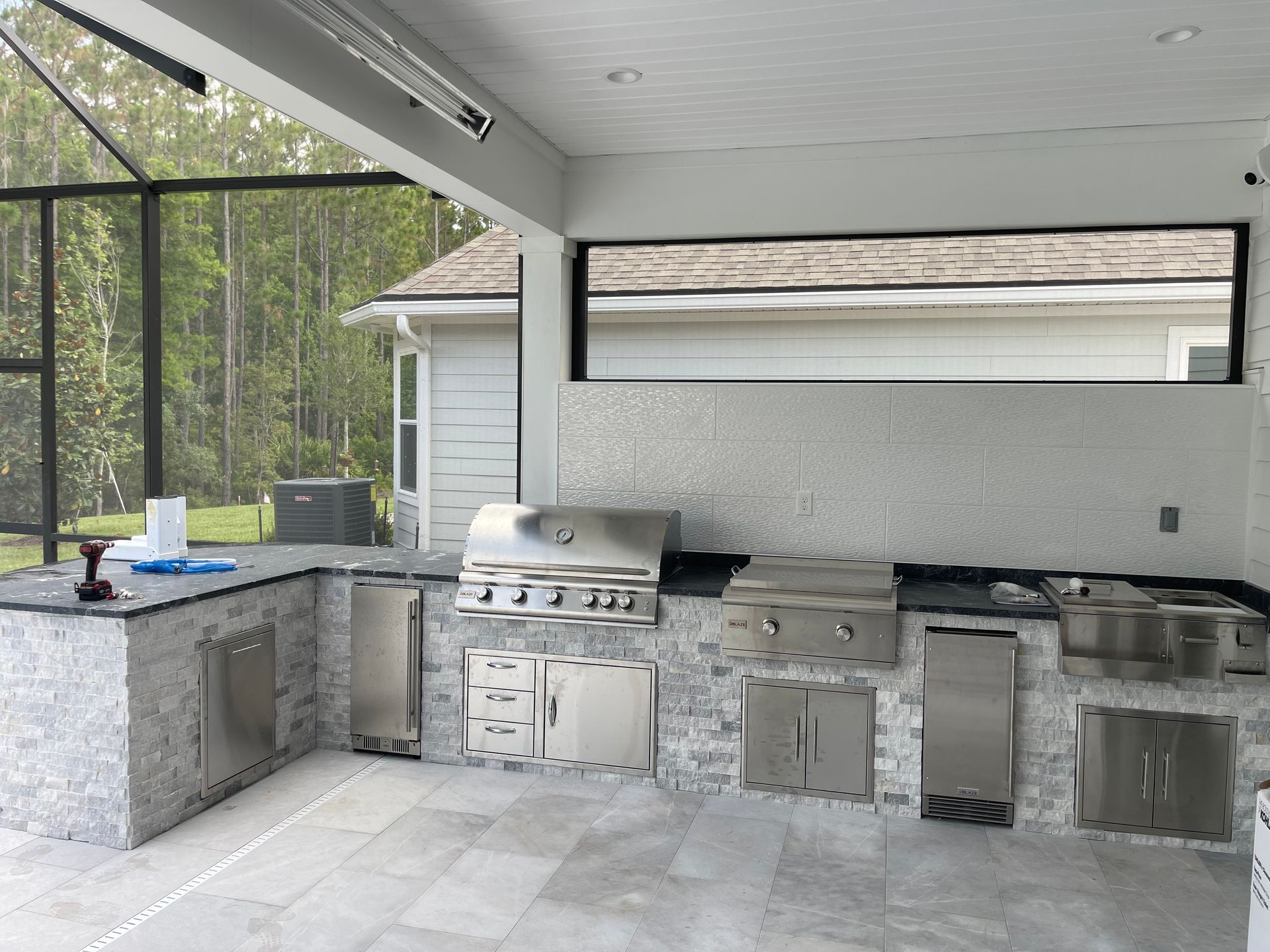 An outdoor kitchen with stainless steel appliances, gray countertops, and a tile backsplash and floor. It's under a covered patio with a screened enclosure.