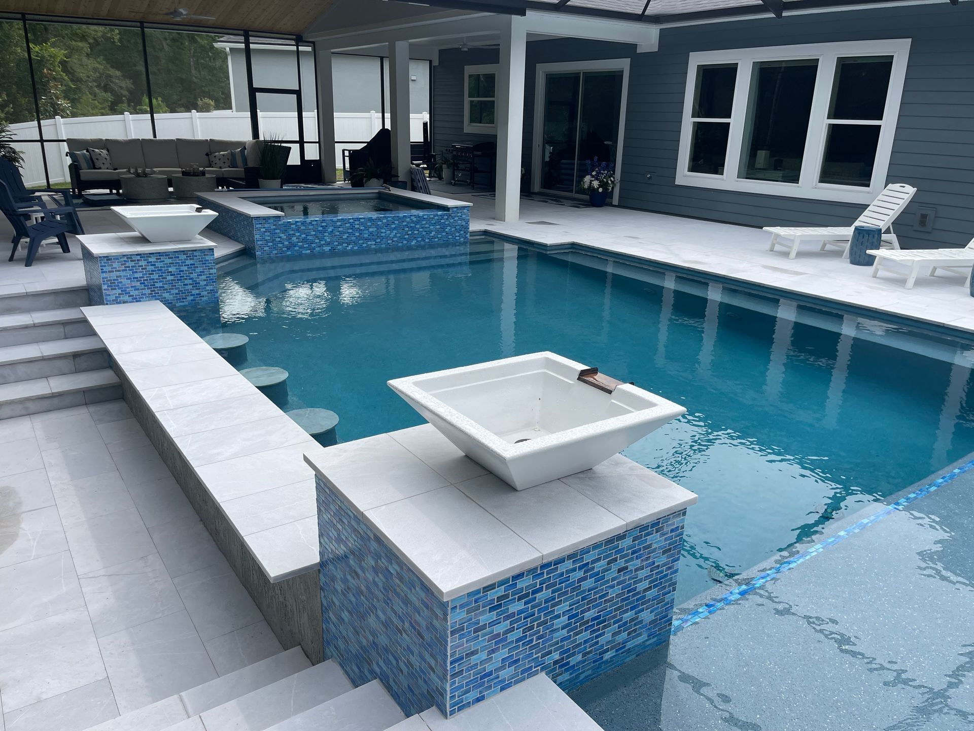 A backyard pool with blue tiled surfaces and a bar area. White and blue tiles surround the pool with steps, a fire pit, and a covered patio.
