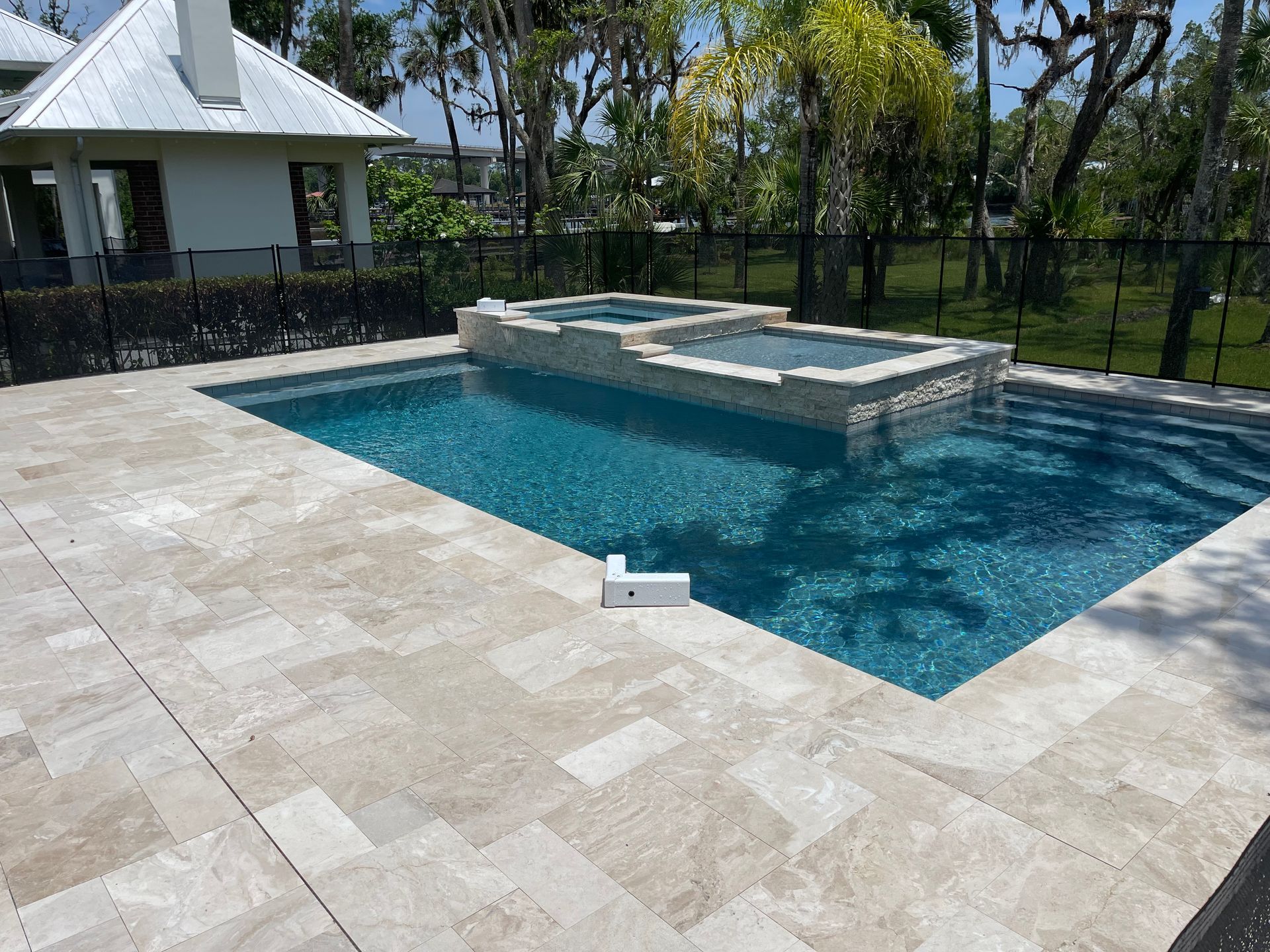 A backyard pool with a spa, surrounded by beige stone tiles. The pool has blue water and is near a white building.