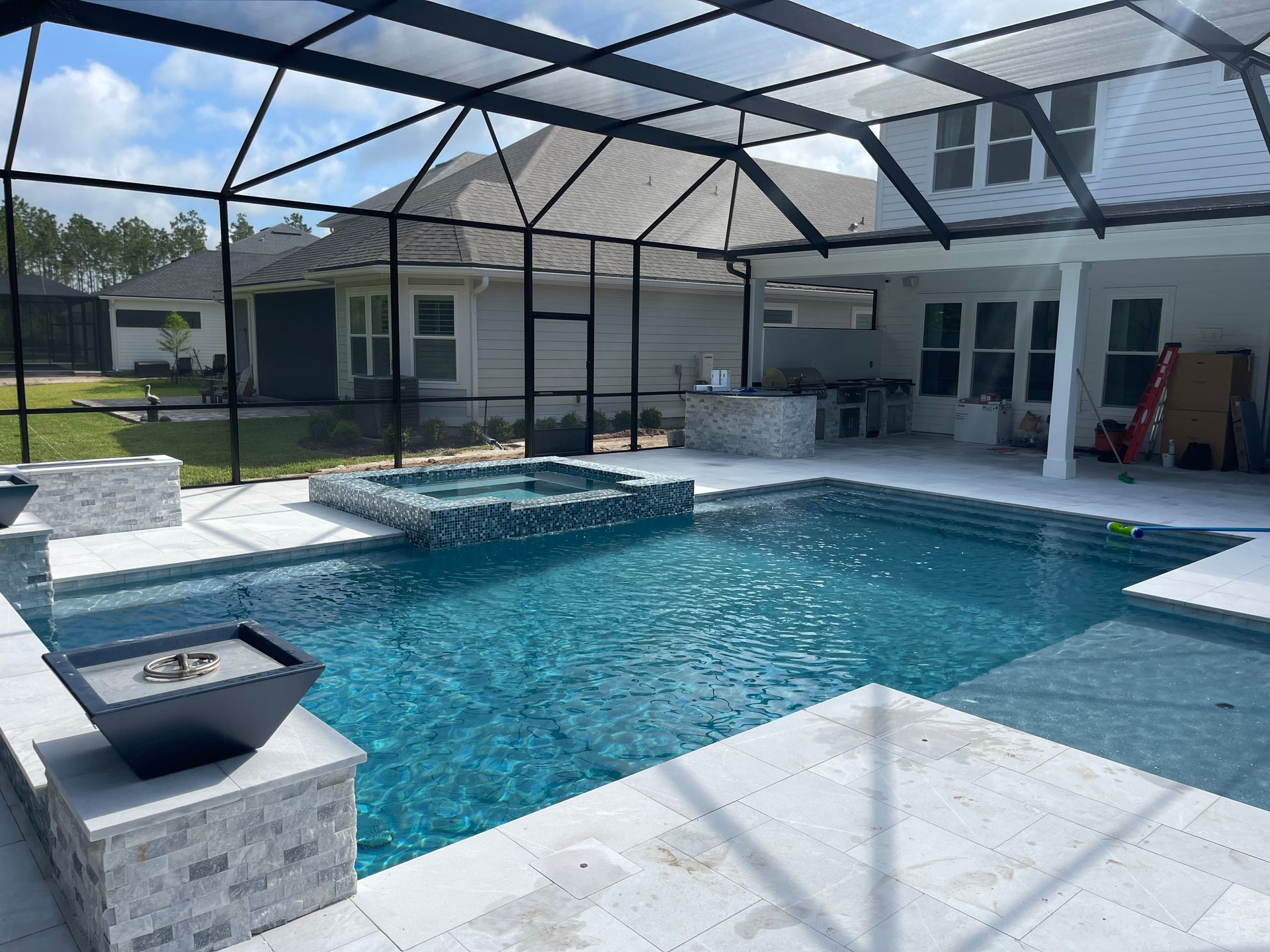 A luxurious pool with a hot tub, a fire pit, and a screened enclosure on a sunny day. The water is blue, and the patio is white.