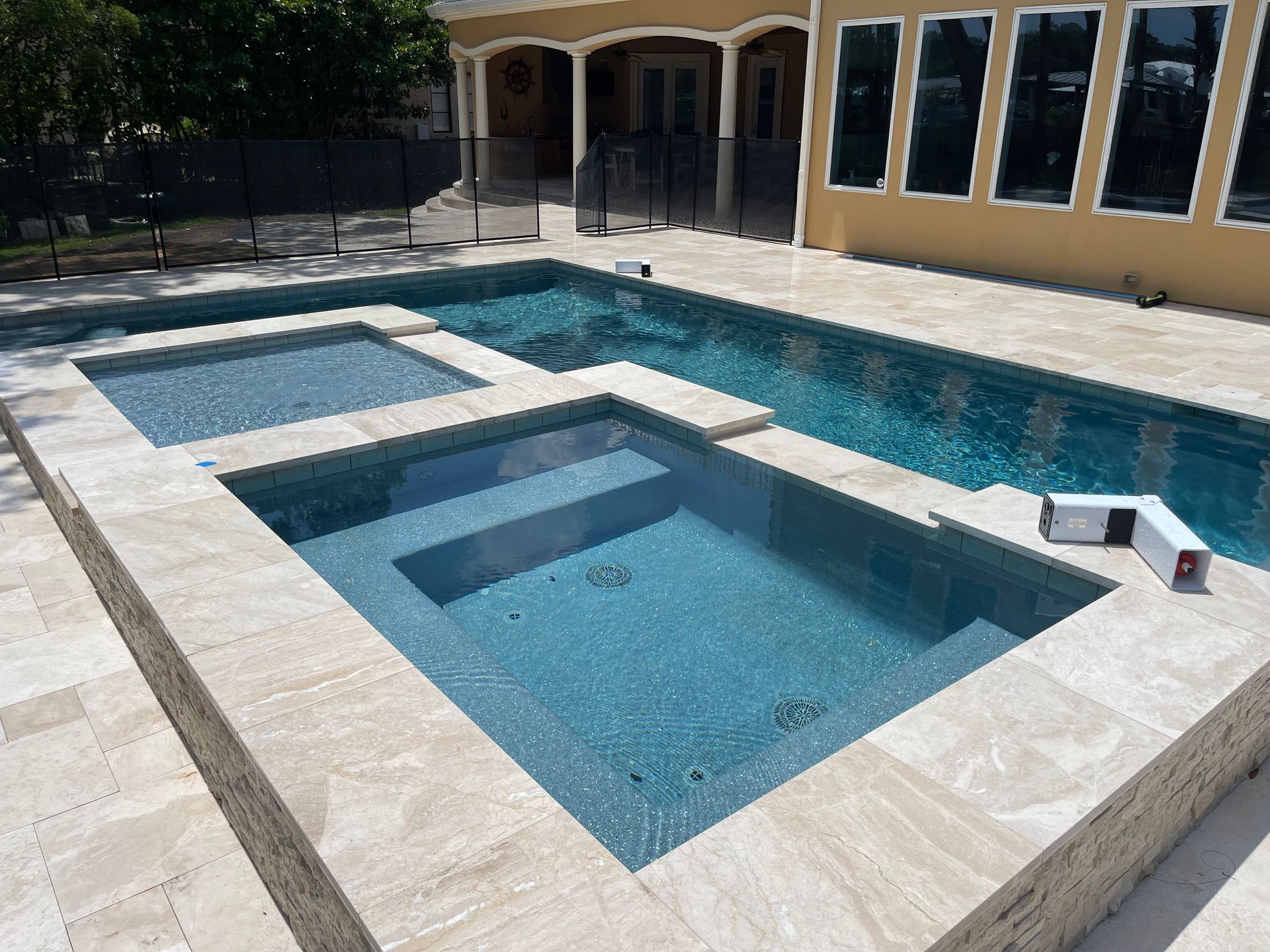 A multi-level swimming pool and spa with blue tile and light stone borders, next to a yellow stucco building.