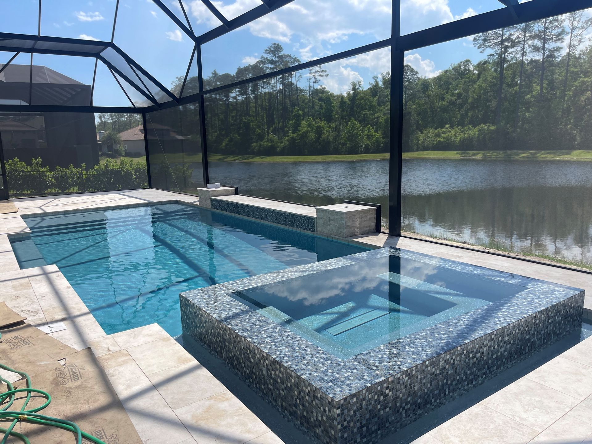 Pool and spa with blue water, surrounded by light-colored stone. A screen enclosure and natural scenery are visible in the background.
