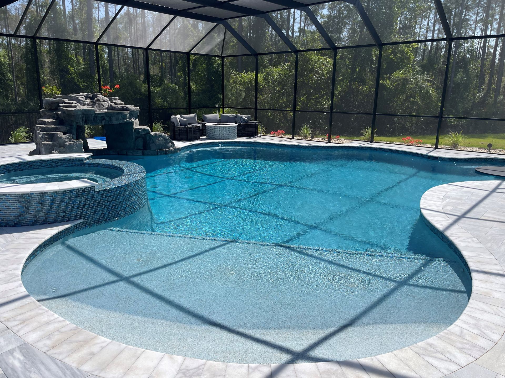 Pool with a waterfall, jacuzzi, and screened enclosure. Light blue water contrasts with gray stone and black screening.