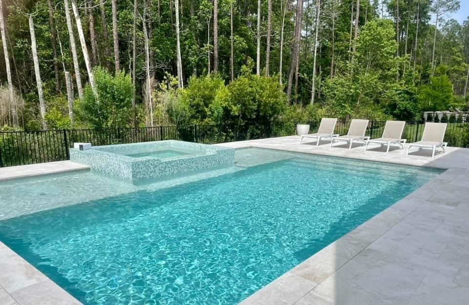 Backyard pool with jacuzzi and multiple lounge chairs, set in a forest. Turquoise water reflects sunlight.