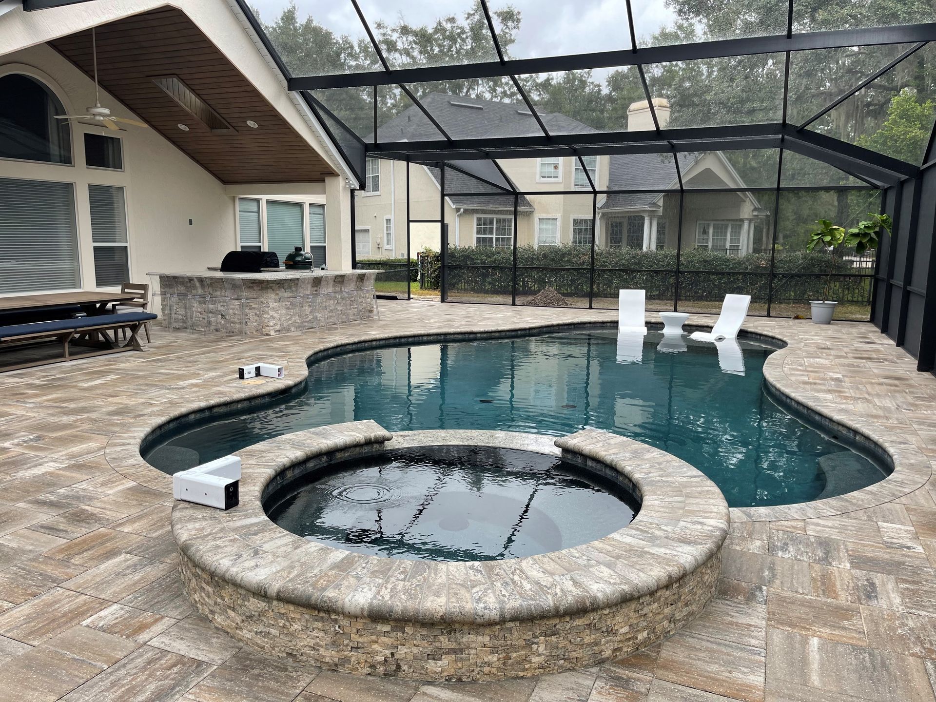 A backyard with a pool and spa enclosed by a screened structure. There's a stone patio, an outdoor kitchen, and white lounge chairs.