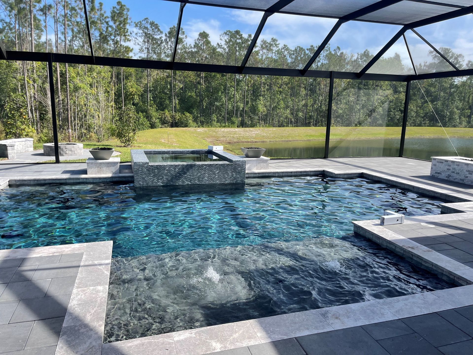 A swimming pool with turquoise water, surrounded by gray stone and screened in, with trees visible in the background.