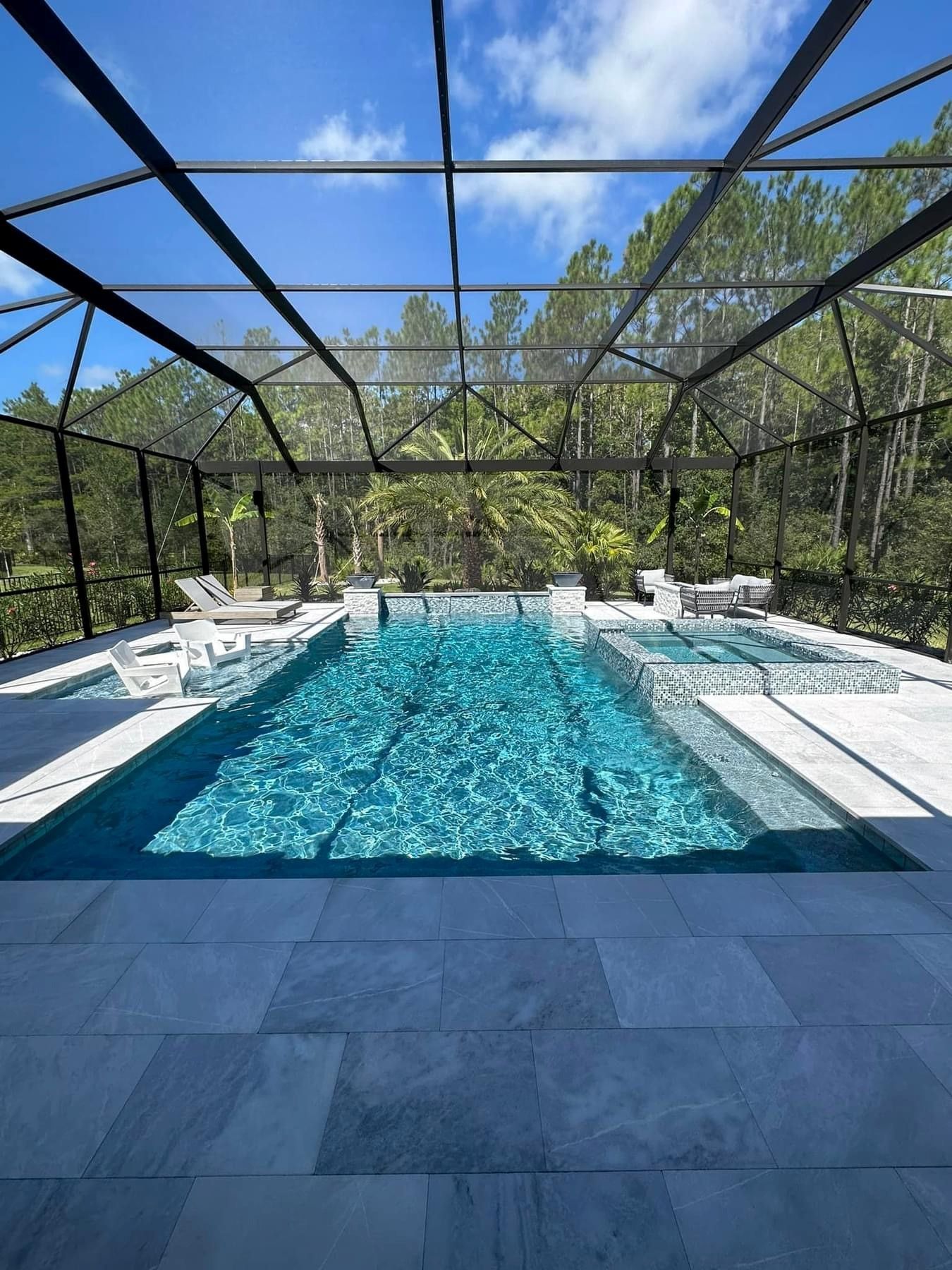 A rectangular pool with clear, teal water enclosed by a black screened structure. White lounge chairs sit on the pool's edge, with a backdrop of trees.