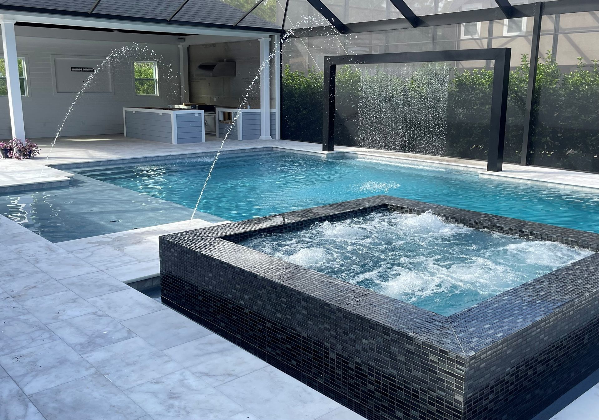 A luxurious backyard pool with a built-in jacuzzi, waterfall, and outdoor kitchen under a covered patio. Blue water reflects sunlight.