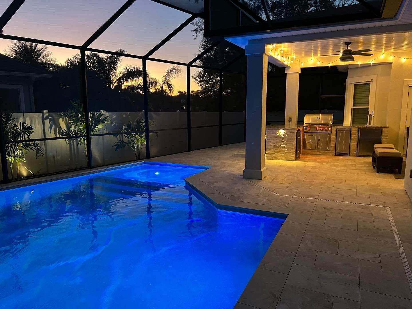 Nighttime outdoor patio and pool area with a glowing blue pool, illuminated grill, and string lights.
