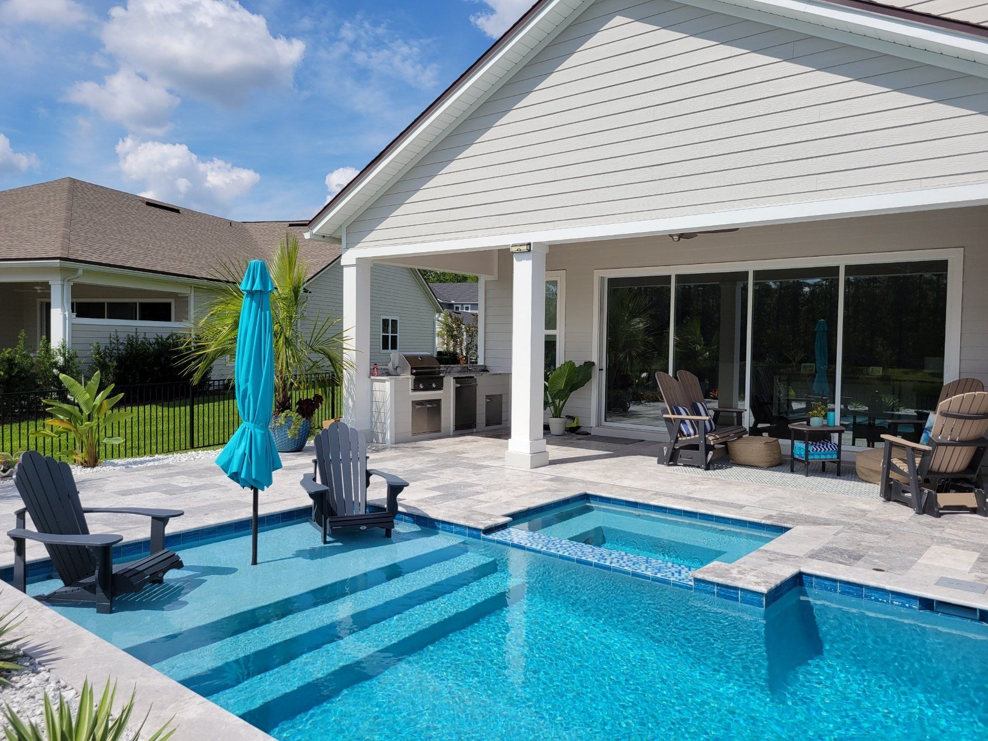 Backyard patio with a blue pool, seating, and a covered grilling area under a light-colored house against a blue sky.