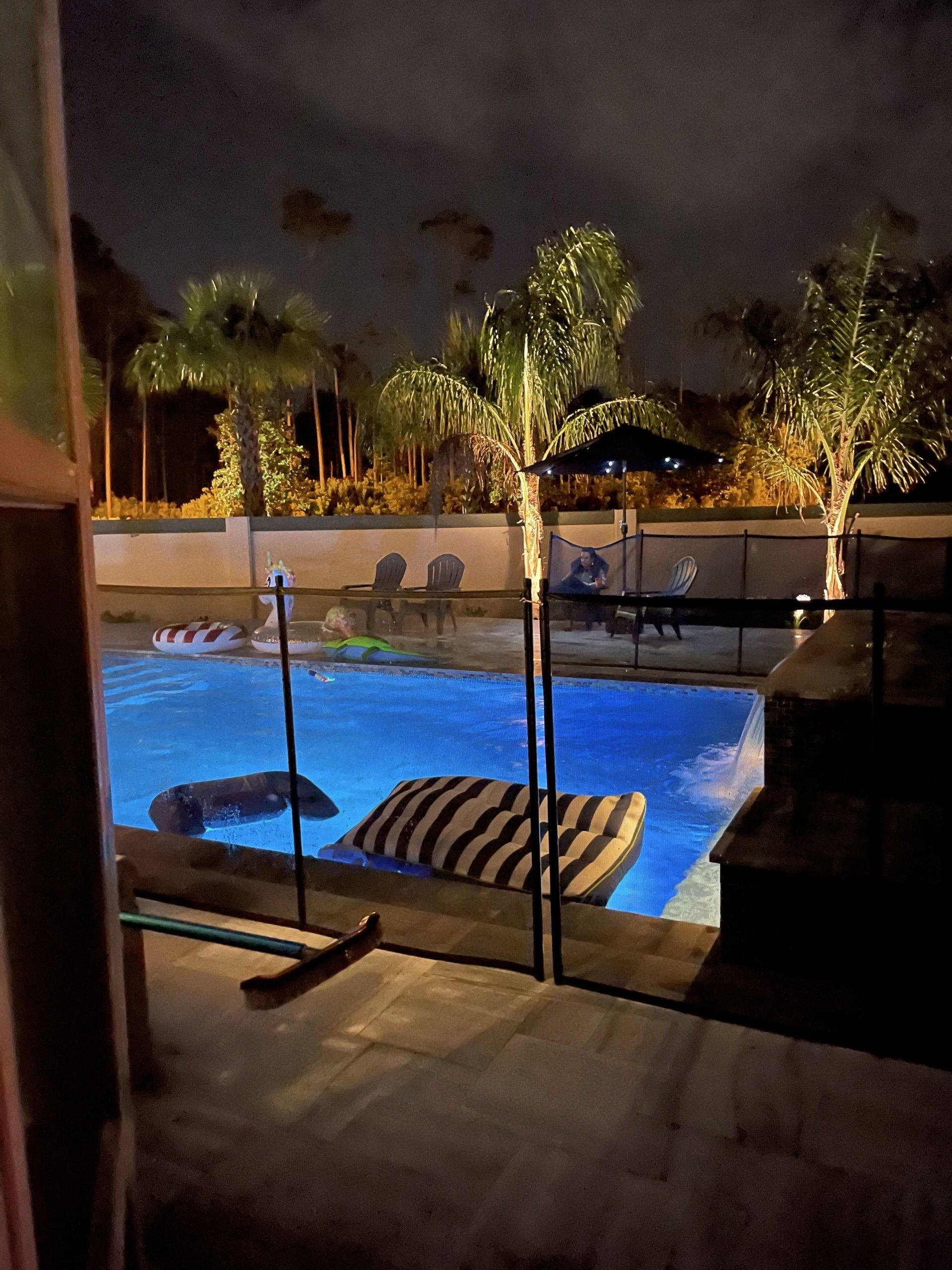 A nighttime view of a pool with illuminated palm trees, filled with floats, and a fence surrounding it.