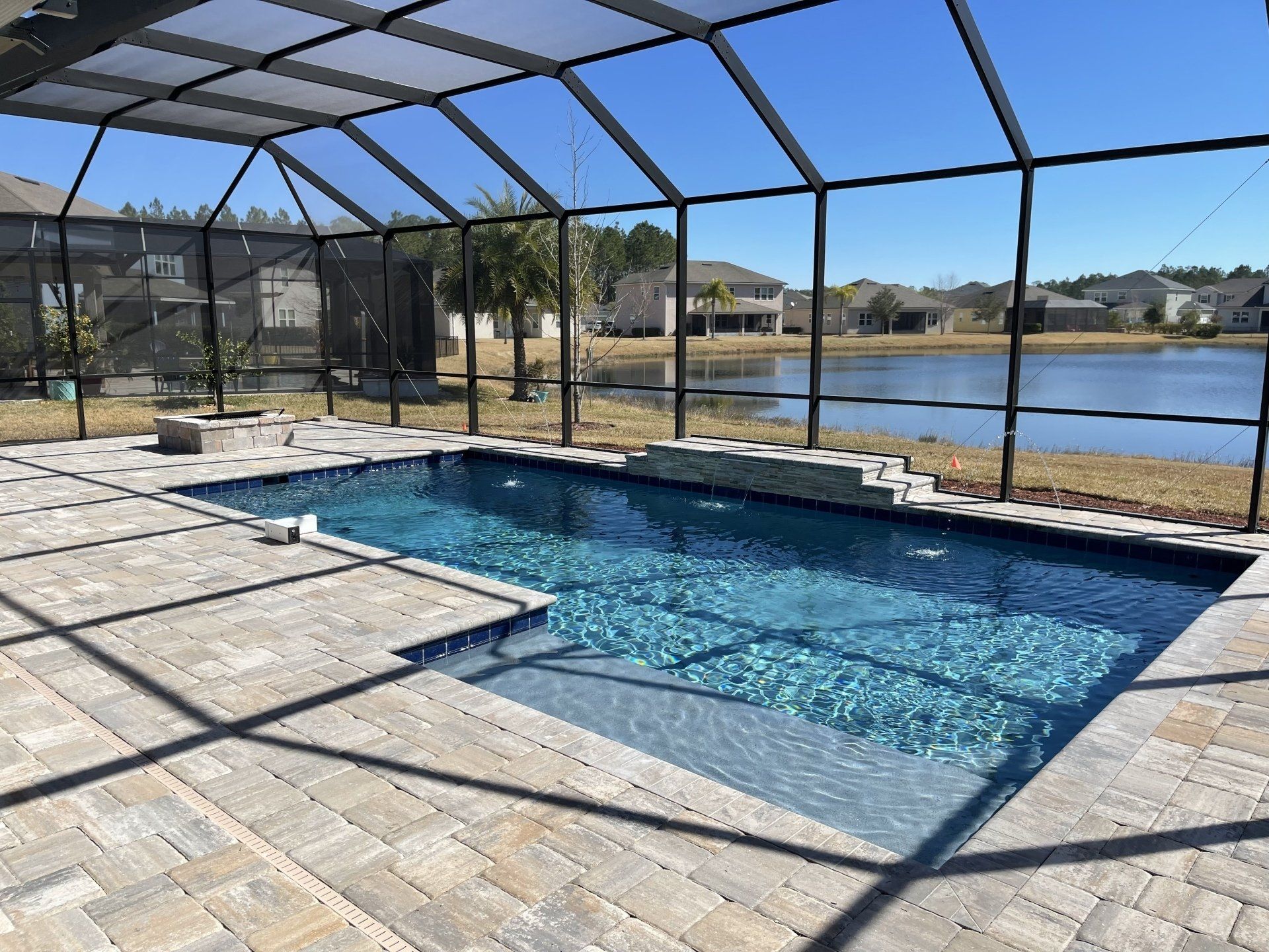 A screened-in rectangular swimming pool with a stone patio overlooking a lake under a clear blue sky.