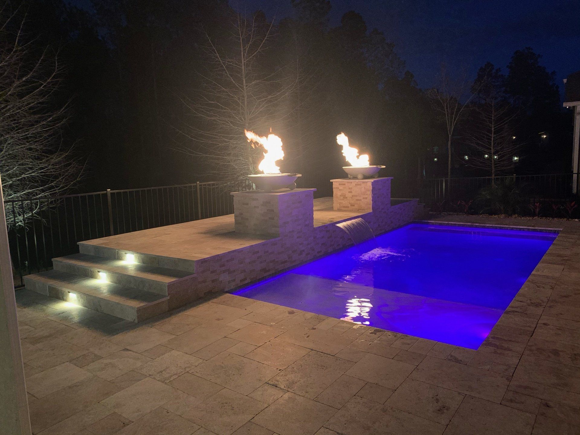 Nighttime pool scene with two fire features burning, stairs with lights, and a pool glowing blue.
