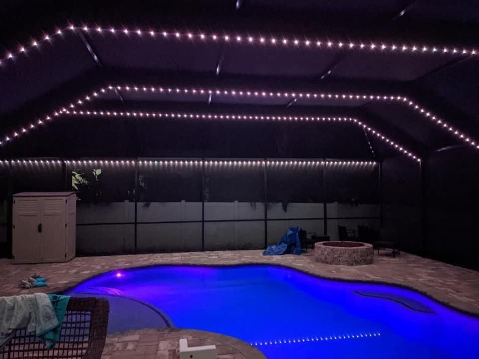 A nighttime pool area with blue-lit water, string lights, and a hot tub. A fire pit sits beside a small slide.