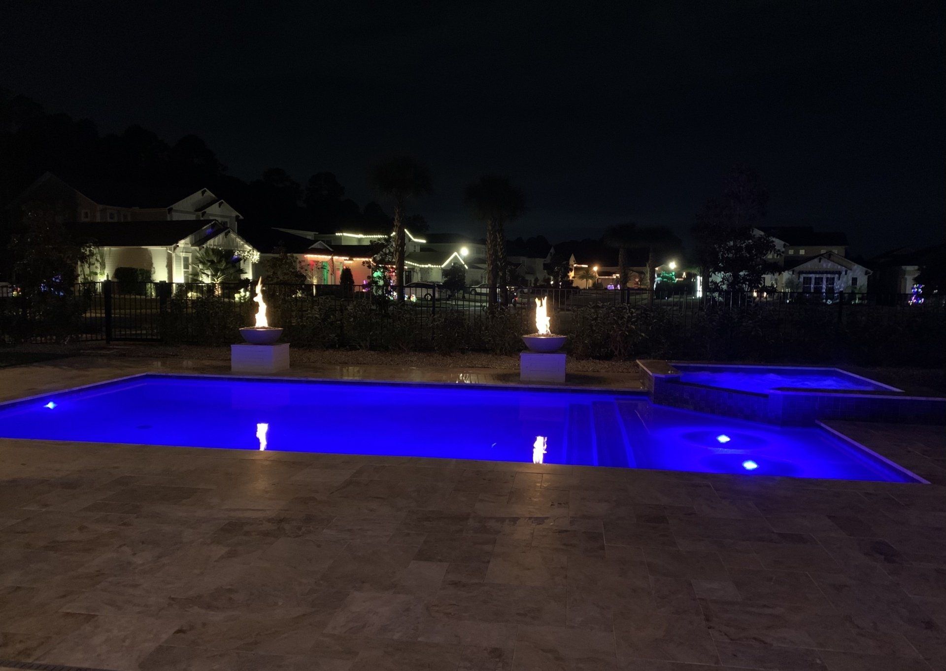 A nighttime shot of a pool with blue lights. Two fire features flank the pool. Several houses are in the background.