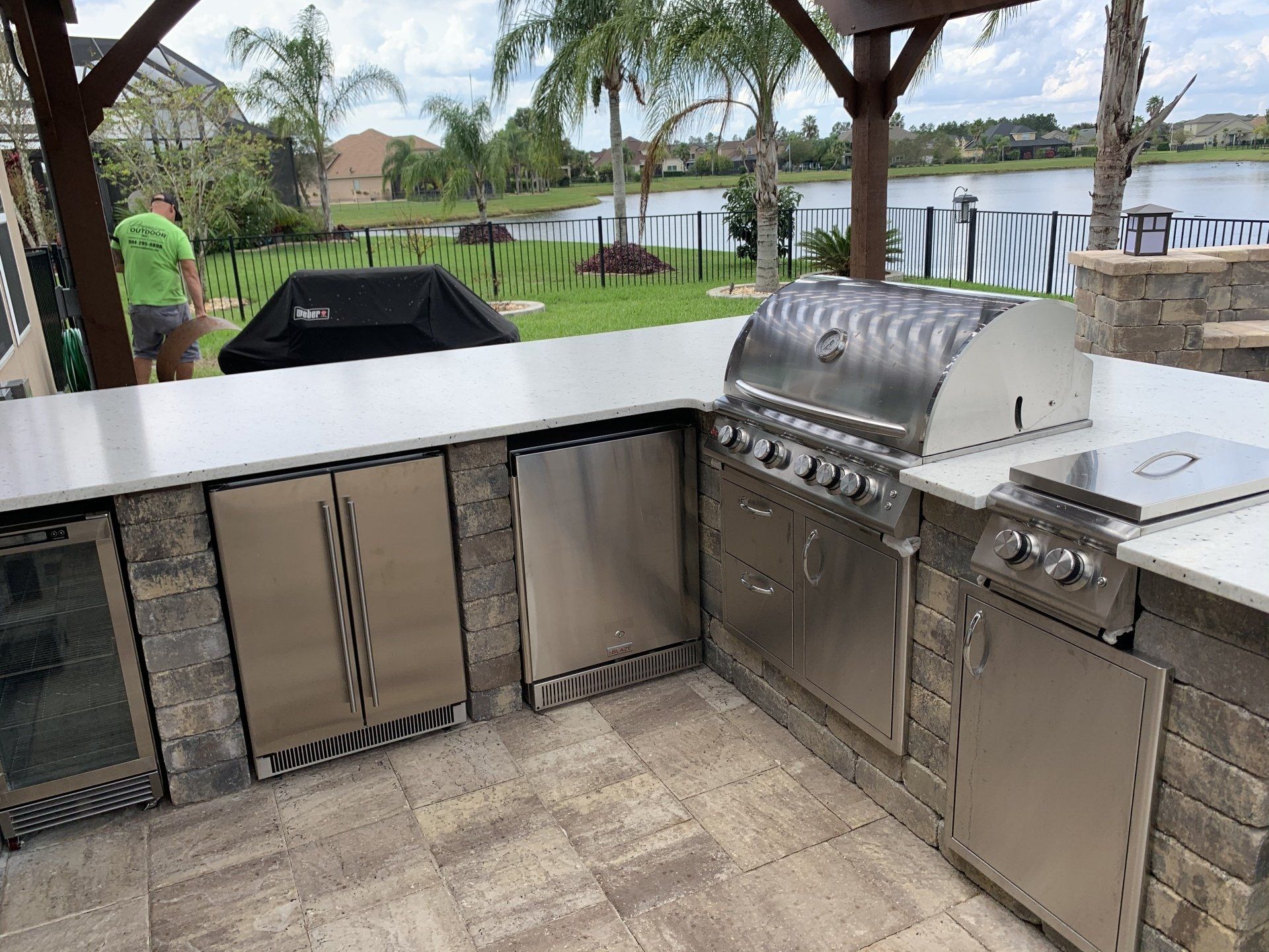 Outdoor kitchen with stainless steel appliances, including a grill and refrigerator, built into a brick structure with a white countertop, overlooking a lake.