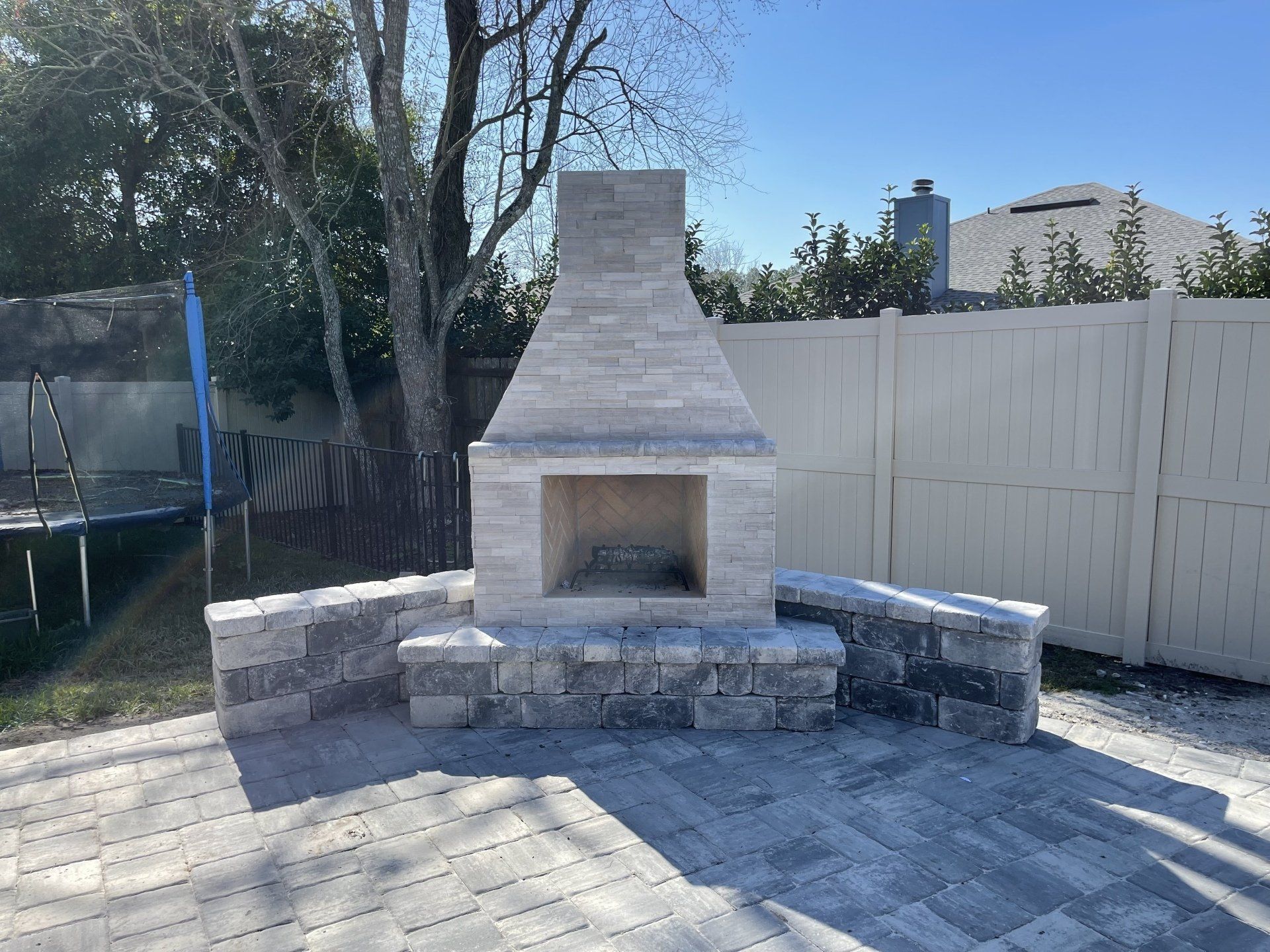 A stone fireplace with a gray brick patio in a backyard setting, with a white fence and a trampoline in the background.