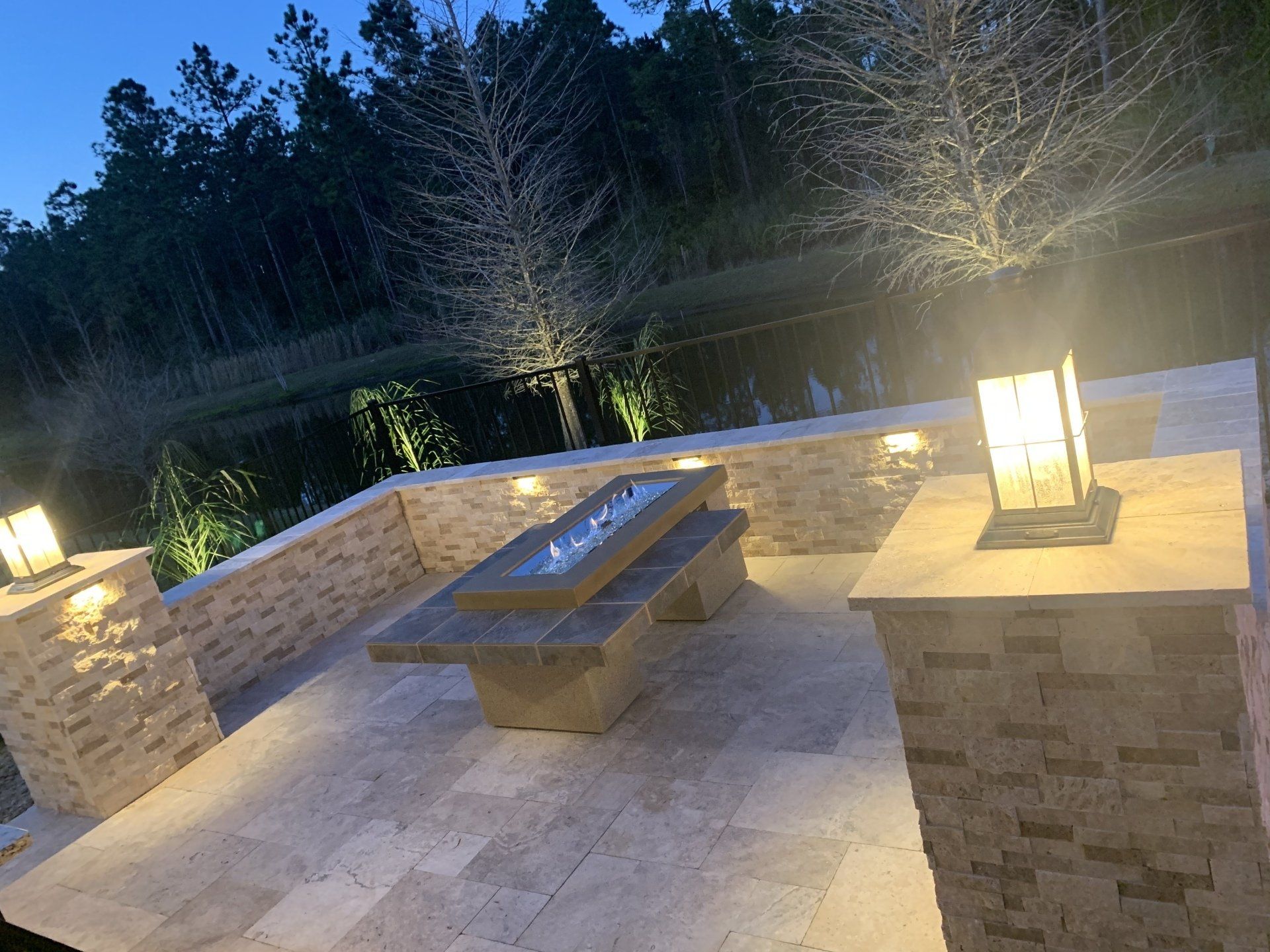 Nighttime patio with a fire pit, lit by warm lights on stone pillars and walls, overlooking a dark lake and trees.
