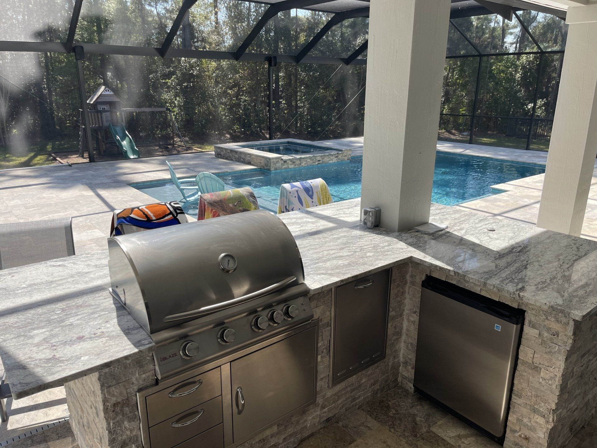 Outdoor kitchen with grill, refrigerator, and countertop overlooking a pool and hot tub. Sunlight and trees visible in the background.