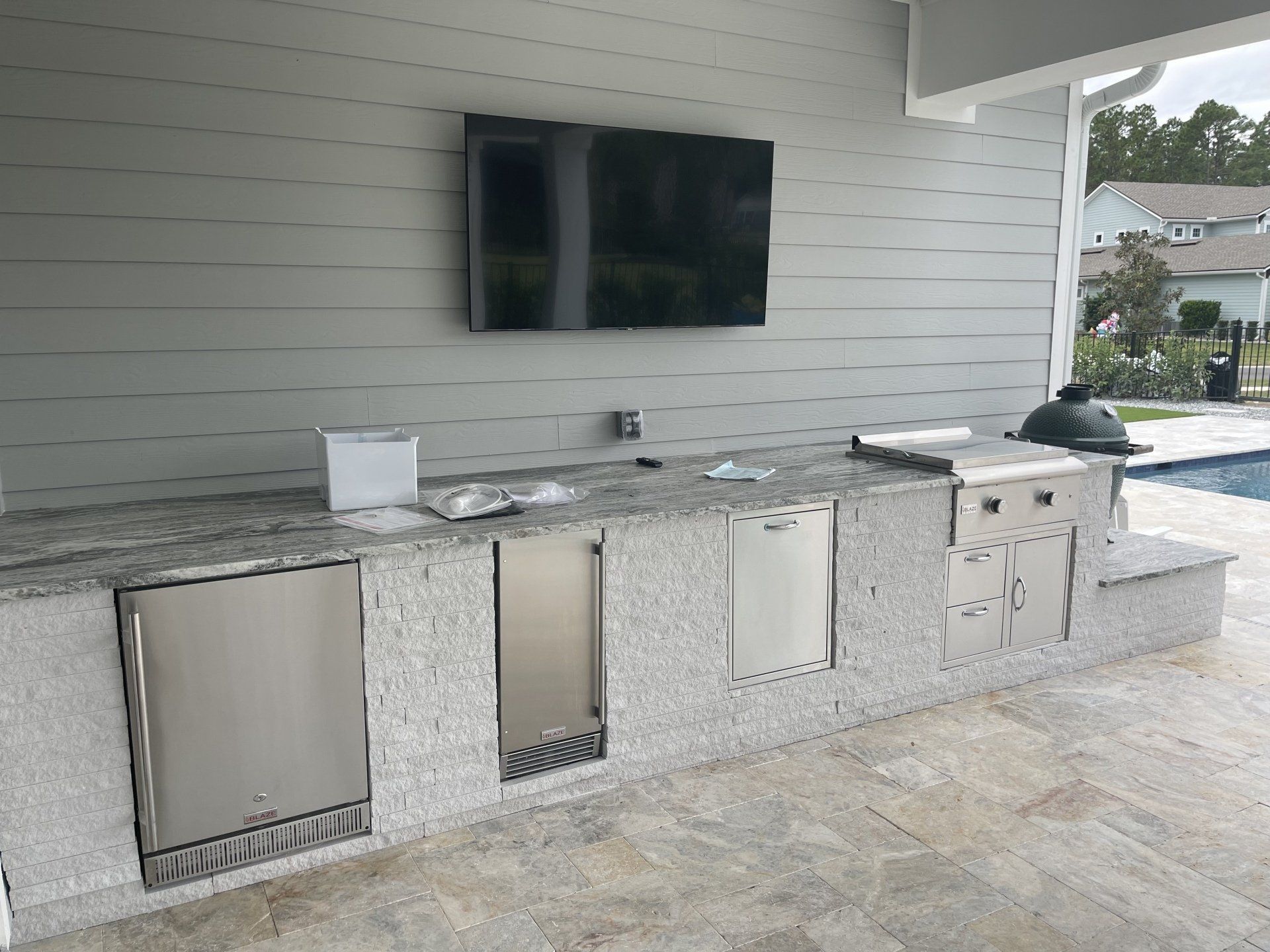 Outdoor kitchen with a granite countertop, grill, refrigerator, and a mounted TV on a light gray wall.