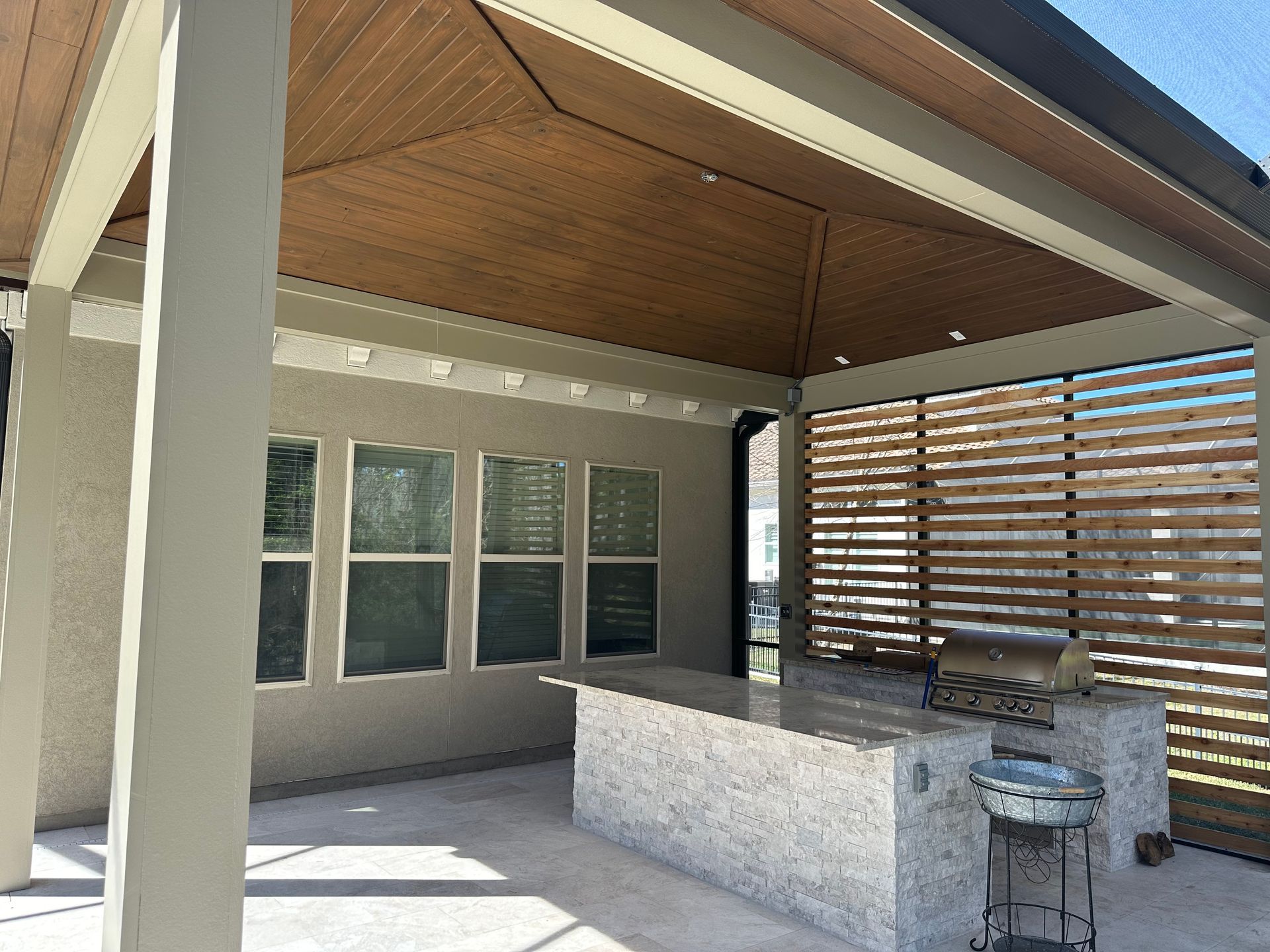Outdoor kitchen with a granite countertop and grill under a wood-paneled ceiling. Features include a slatted wood screen and concrete pillars.