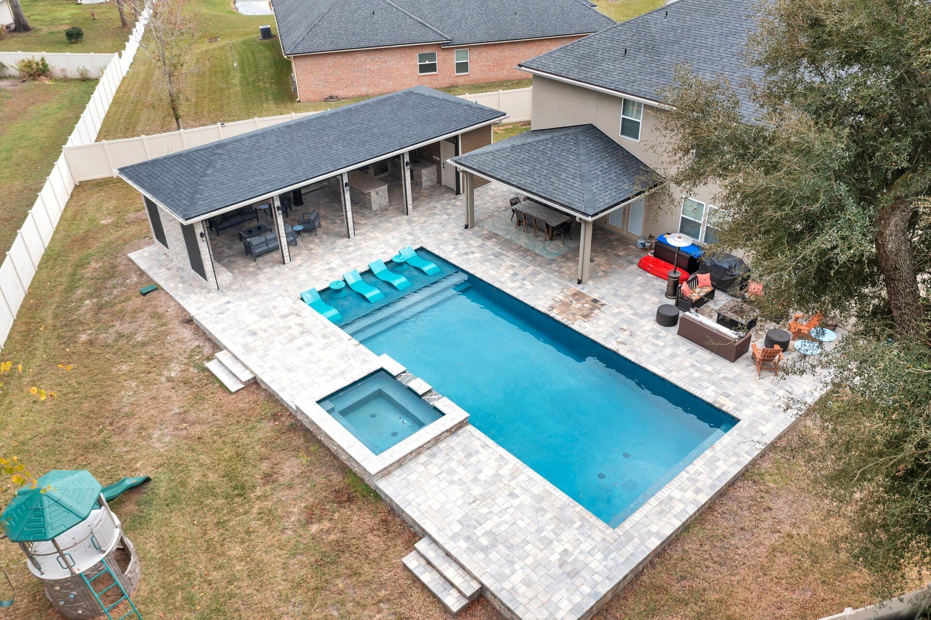 Aerial view of a backyard with a rectangular pool, hot tub, and covered seating areas. There's also a playhouse and outdoor furniture.