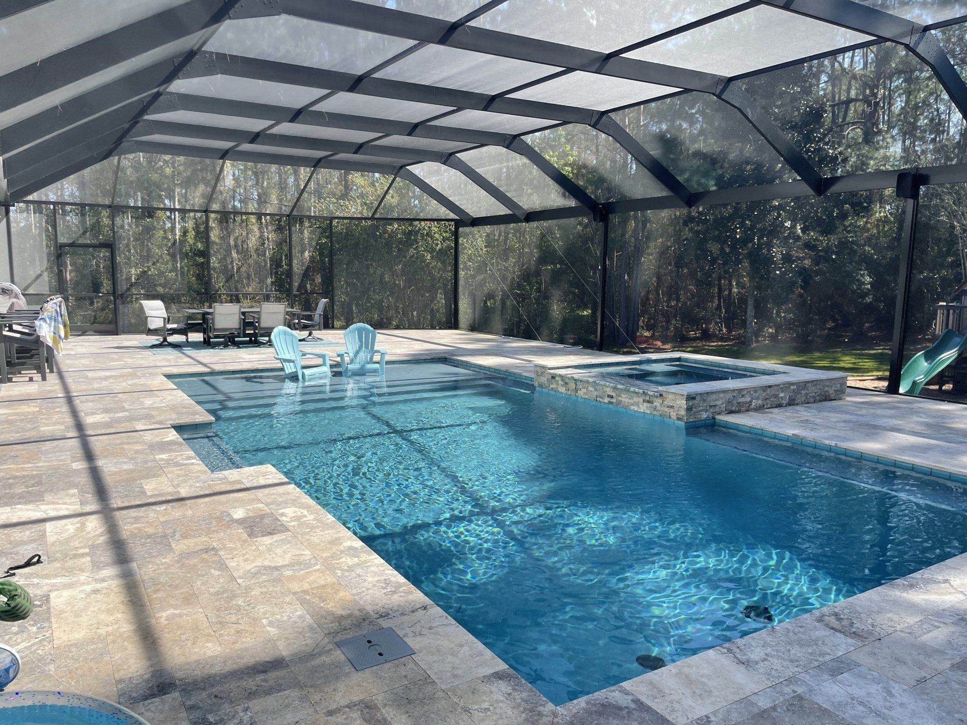 A screened-in pool area with a blue pool, hot tub, and patio furniture. The setting is outdoors with trees visible in the background.