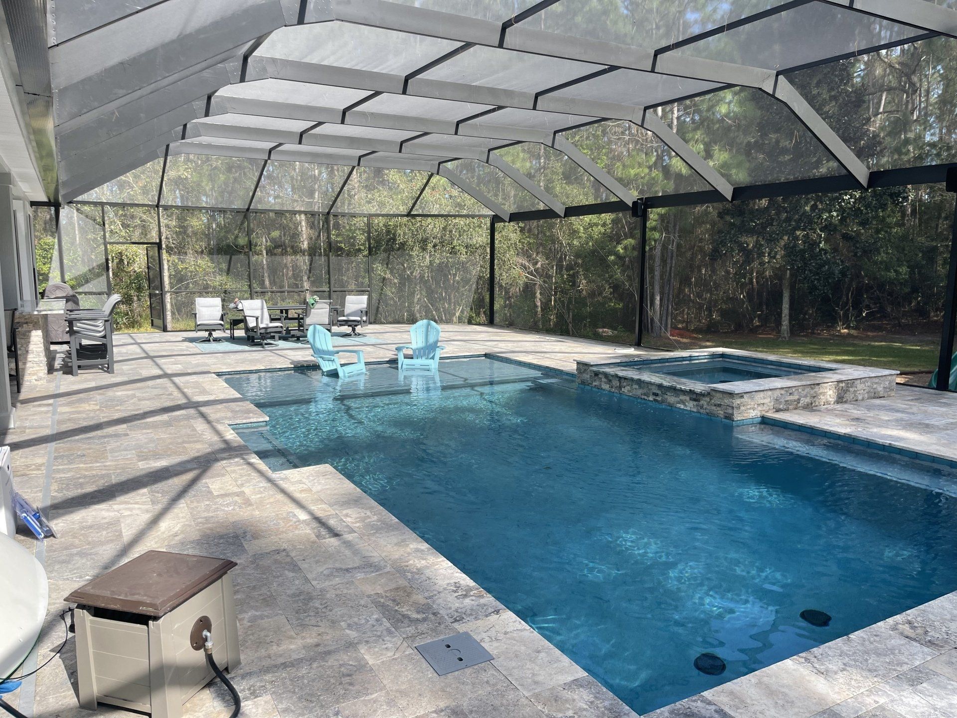 Pool area with a screened enclosure, pool, spa, and seating. Gray pavers, blue water, black screening, and light blue chairs.