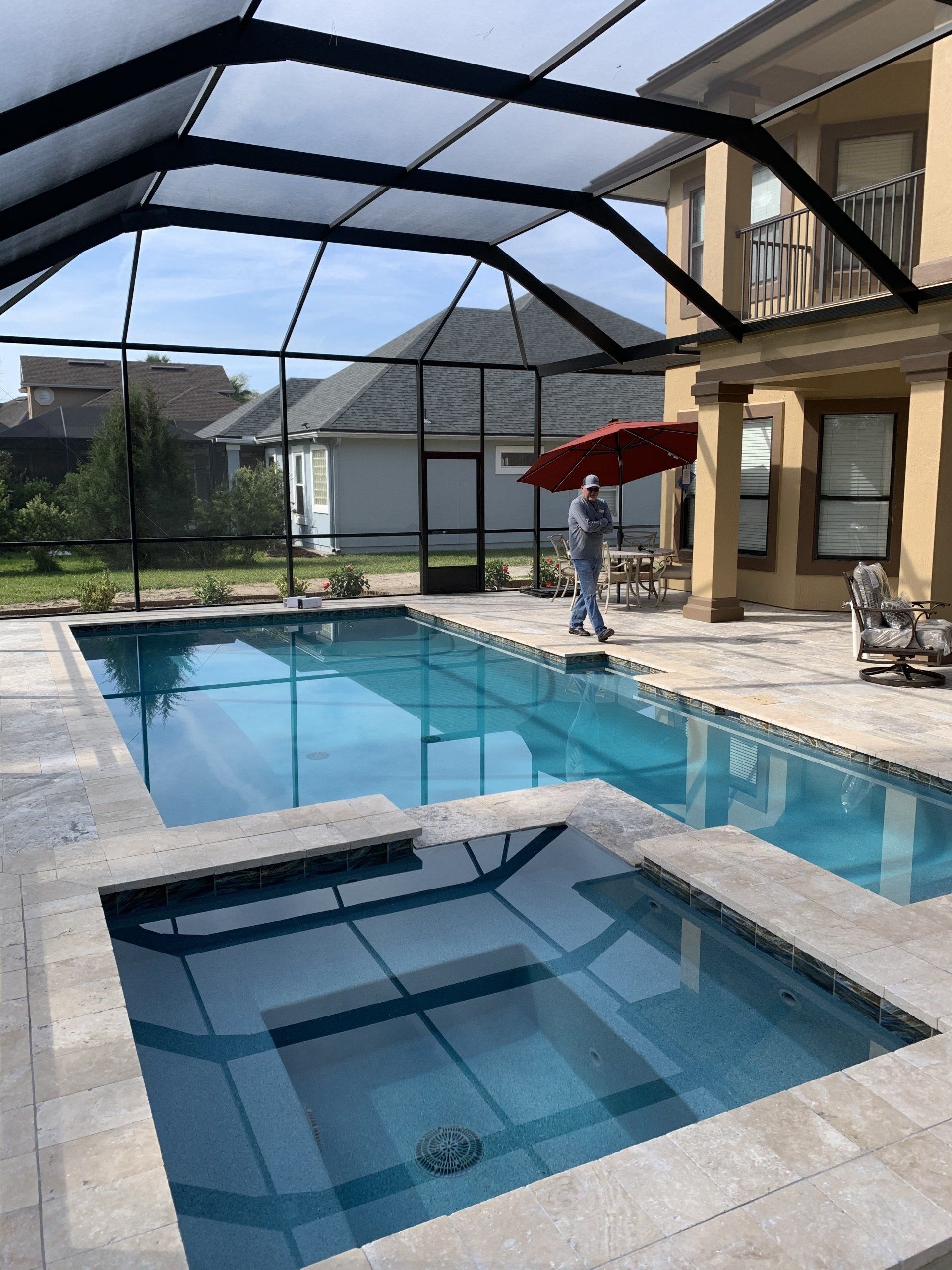 Pool and spa with a covered patio, enclosed by black screening, and a person holding a red umbrella.