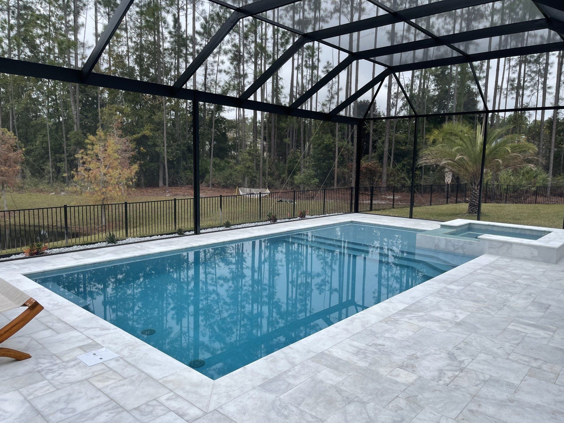 A rectangular swimming pool with a spa, surrounded by light-colored stone. A screened enclosure covers the pool, set against a forested backdrop.