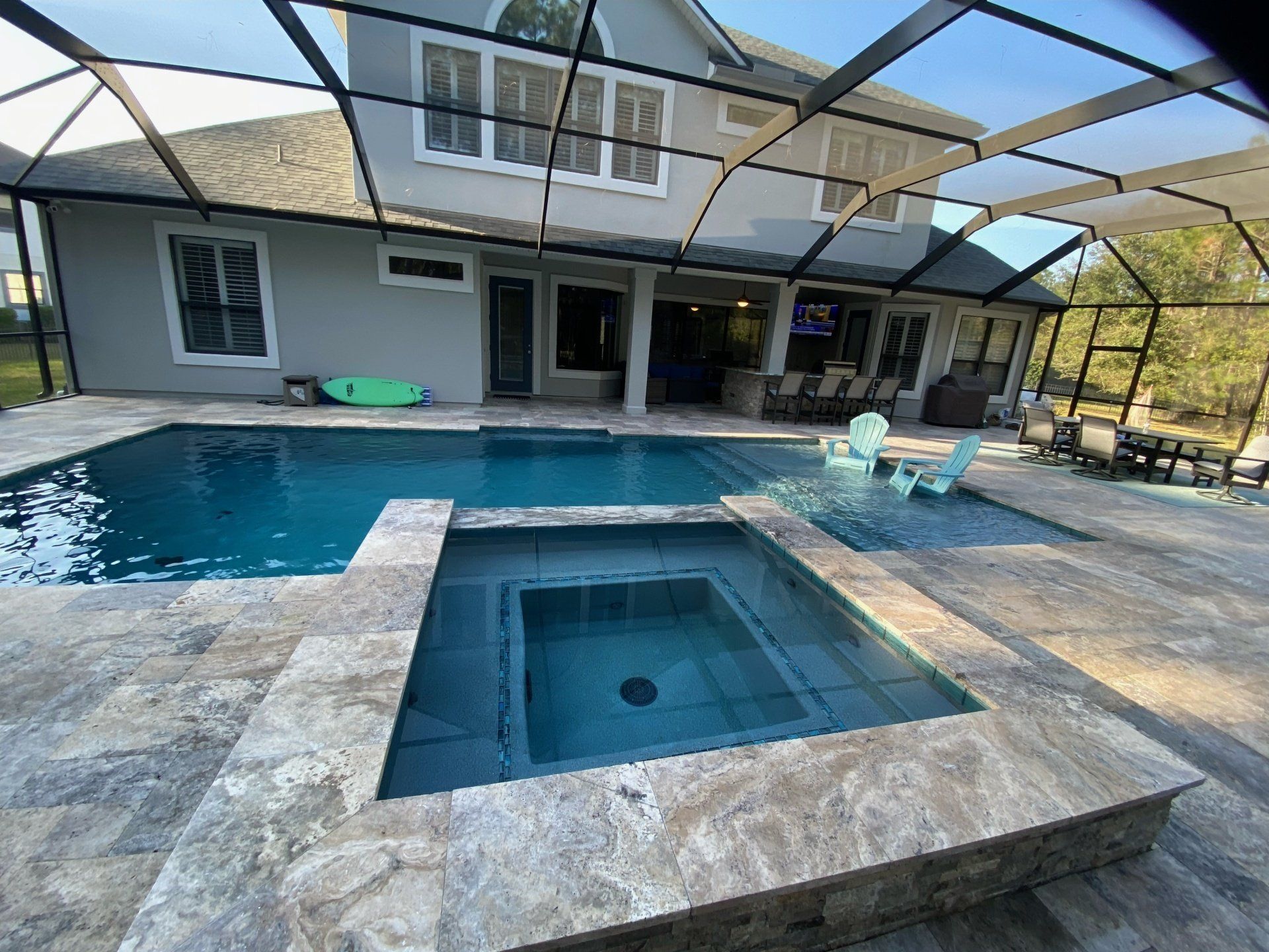 A backyard pool with a spa, patio furniture, and a large house in the background. The pool water is blue, and the day is sunny.