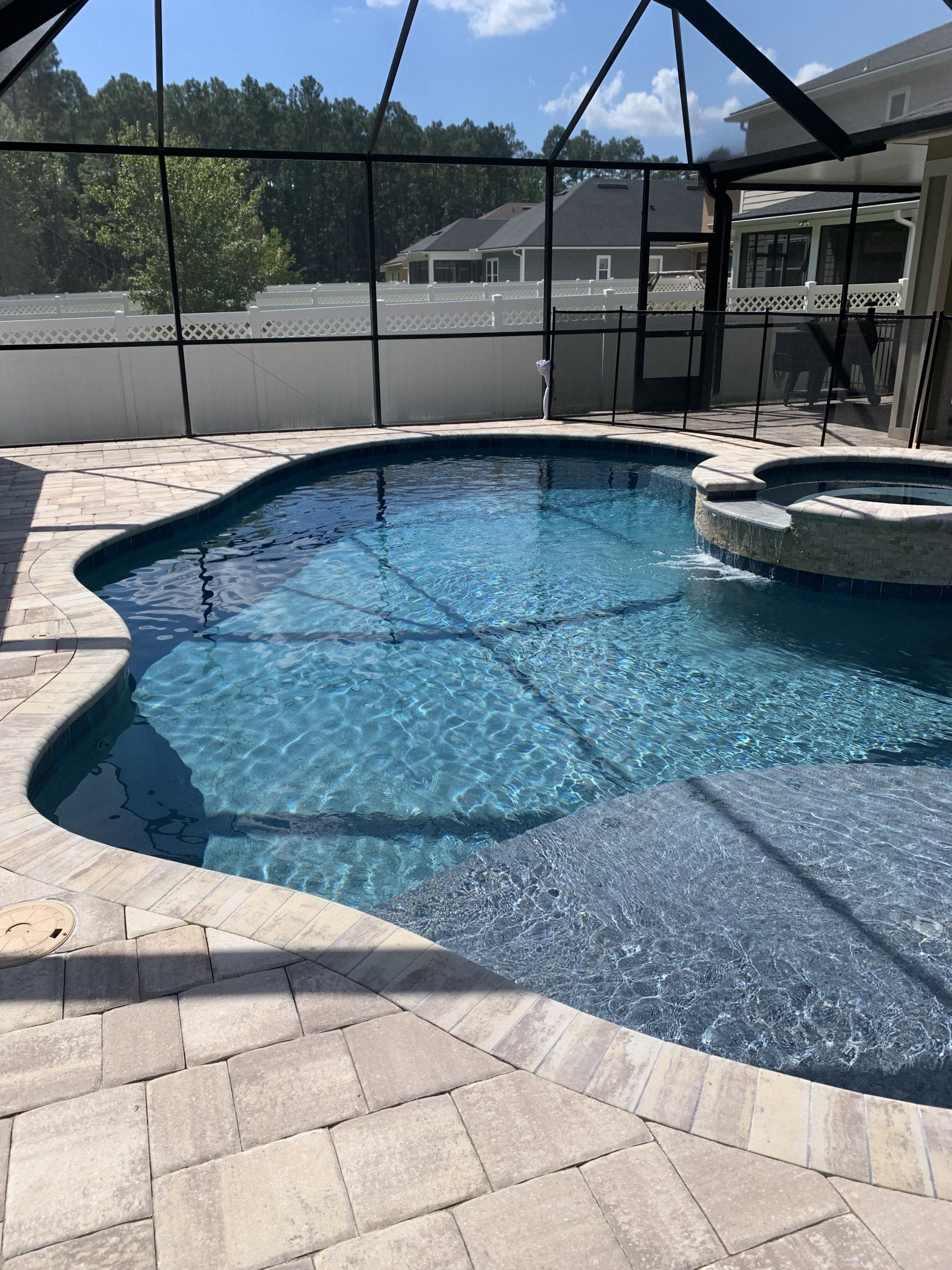 A blue swimming pool with a built-in hot tub, surrounded by a paved patio and a screened enclosure, in an outdoor setting.