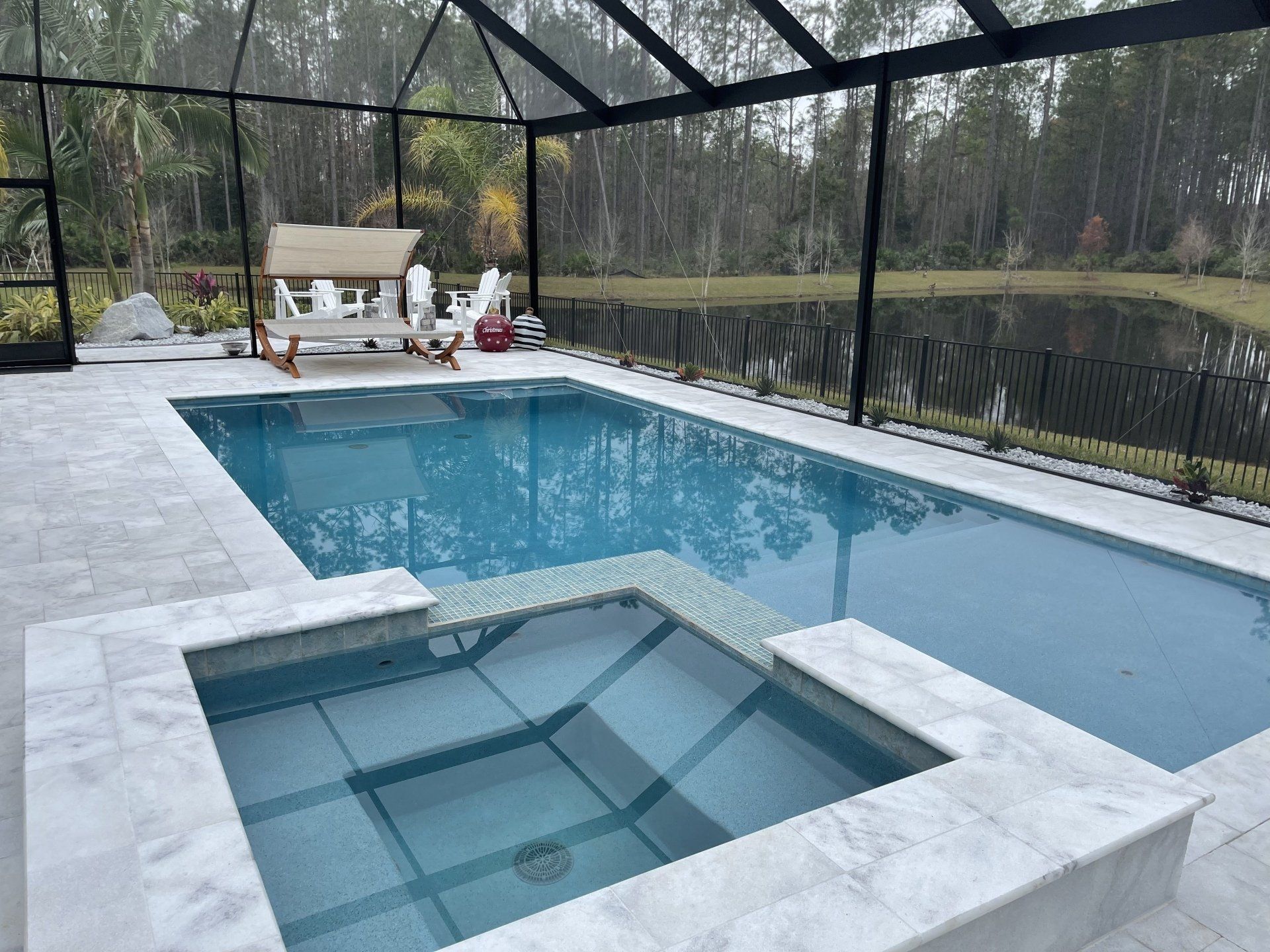 A rectangular swimming pool and spa surrounded by gray tiling, with a screen enclosure and a view of a pond and trees.