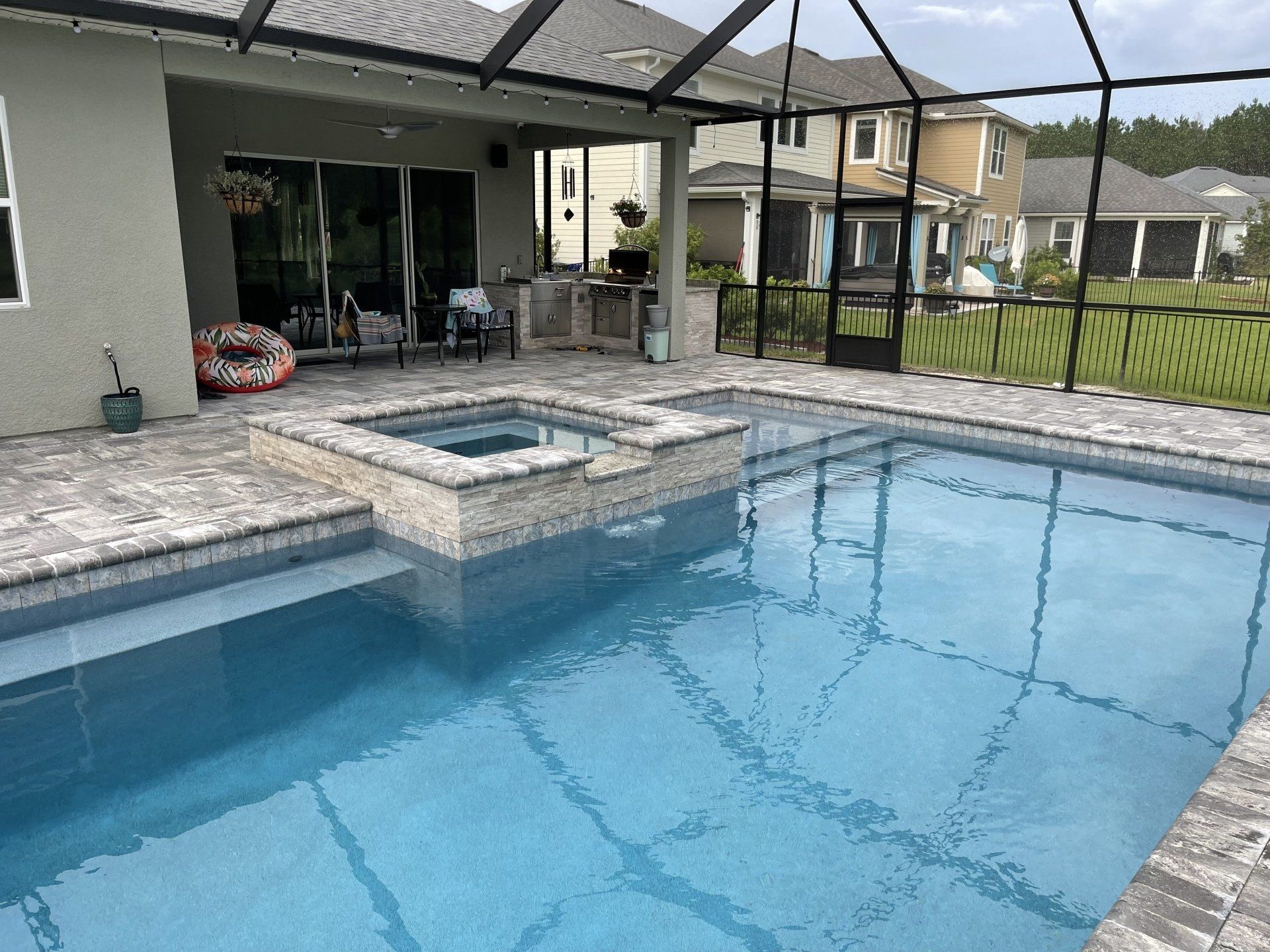 Pool and hot tub with stone surround in a backyard. Screened-in porch, houses, and fence in the background.
