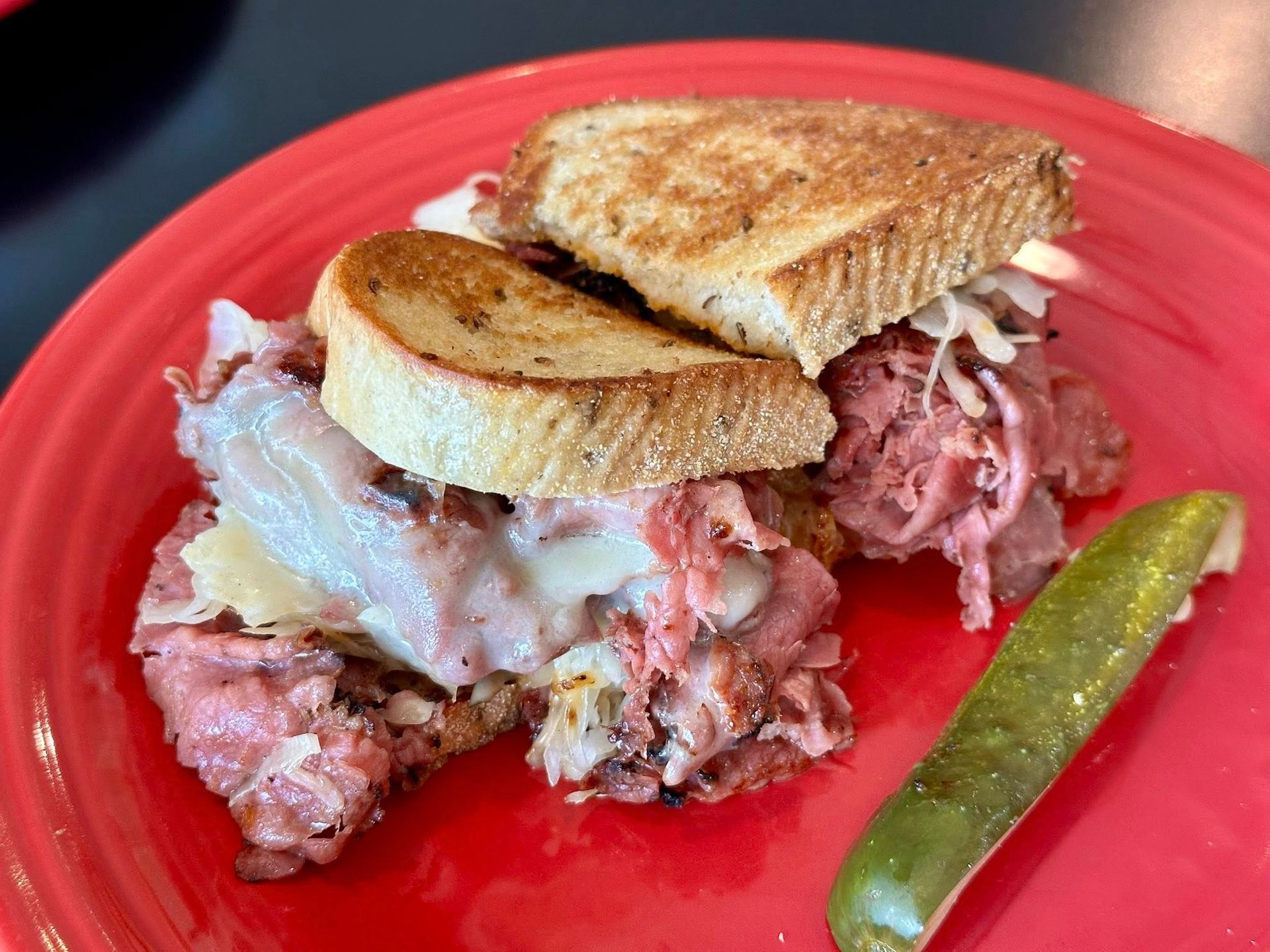 Reuben sandwich on a red plate with a pickle. Toasted bread, corned beef, Swiss cheese, and sauerkraut.