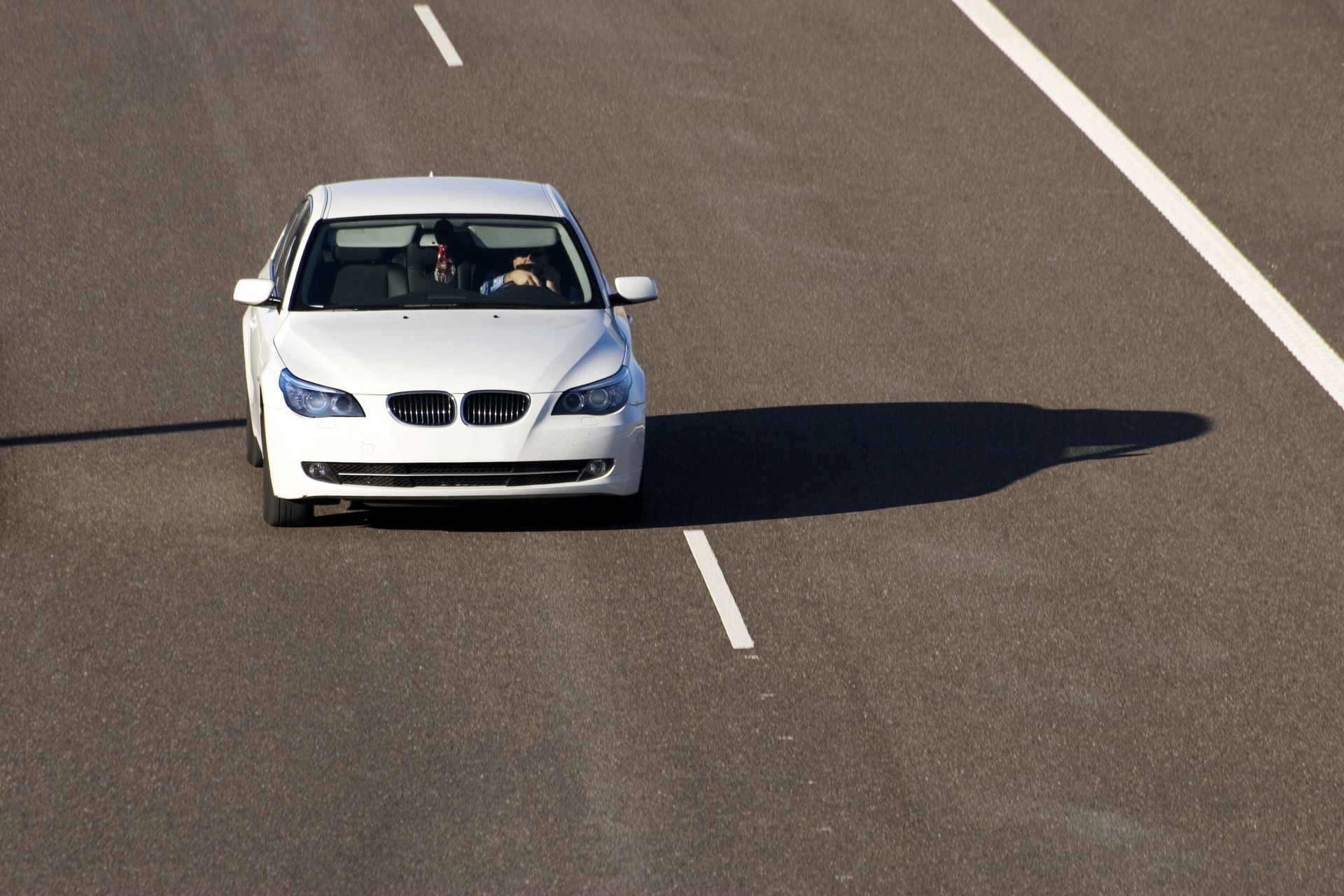 White sedan driving on a highway, centered in the lane with a driver visible inside.