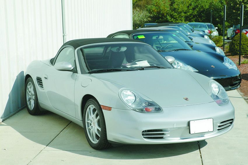 Silver Porsche convertible parked beside a white wall, with blue cars lined up behind it.