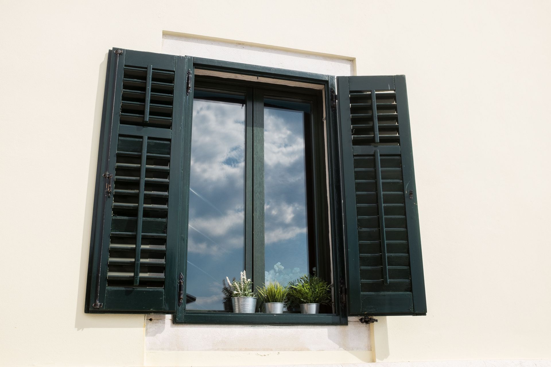 Black shuttered window with cloudy sky reflection and small plants on the sill