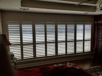 Large window with white shutters letting light into a dim living room with a couch and lamp