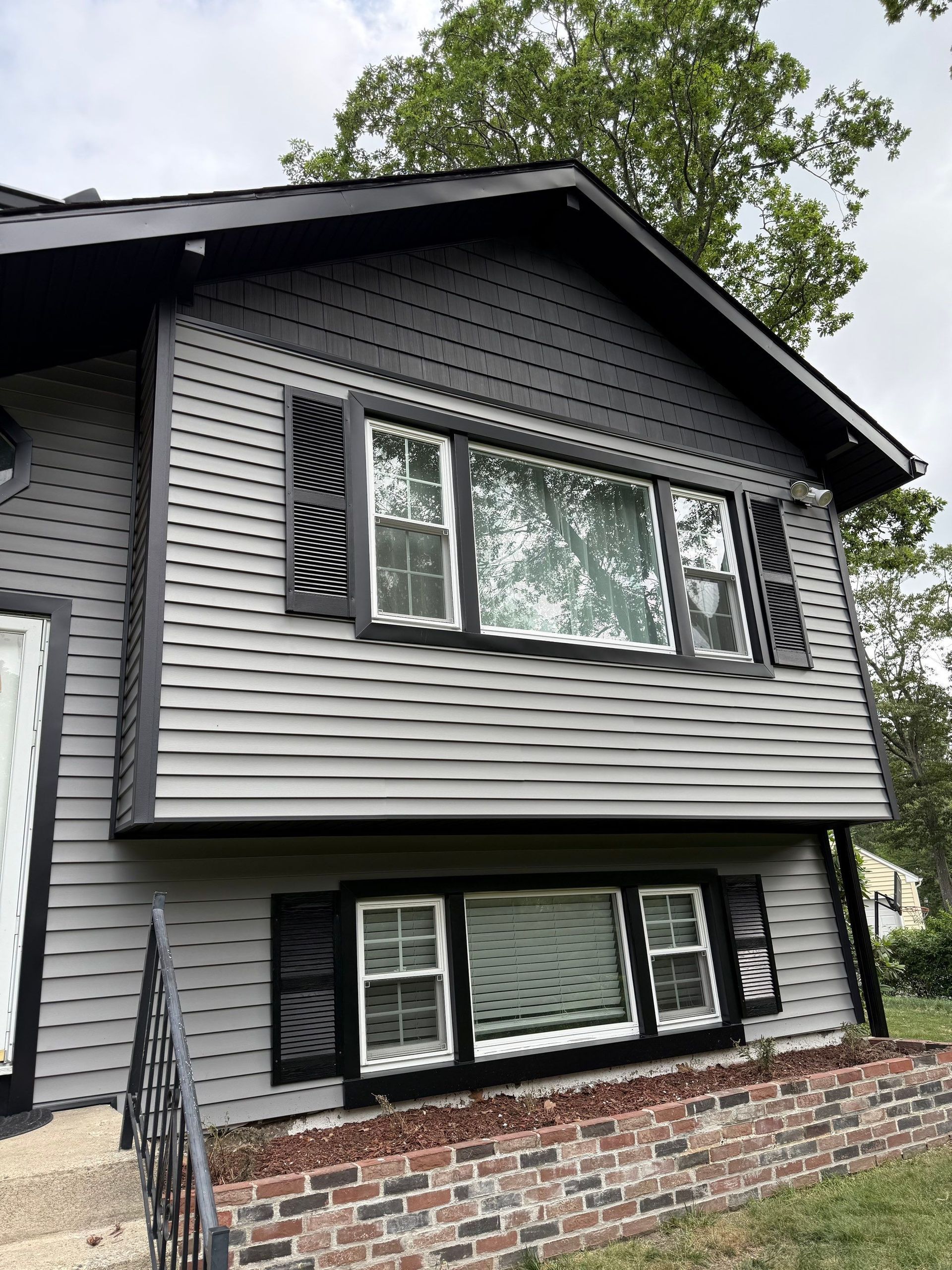 Two-story gray house with black trim and brick lower facade, viewed from outside with trees above