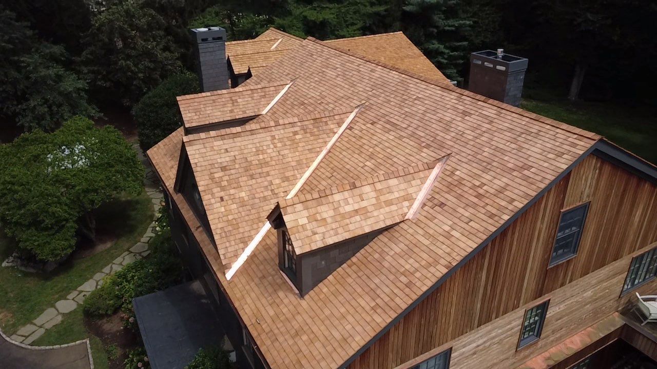 Aerial view of a wooden house with a tan shingle roof and chimneys, surrounded by trees