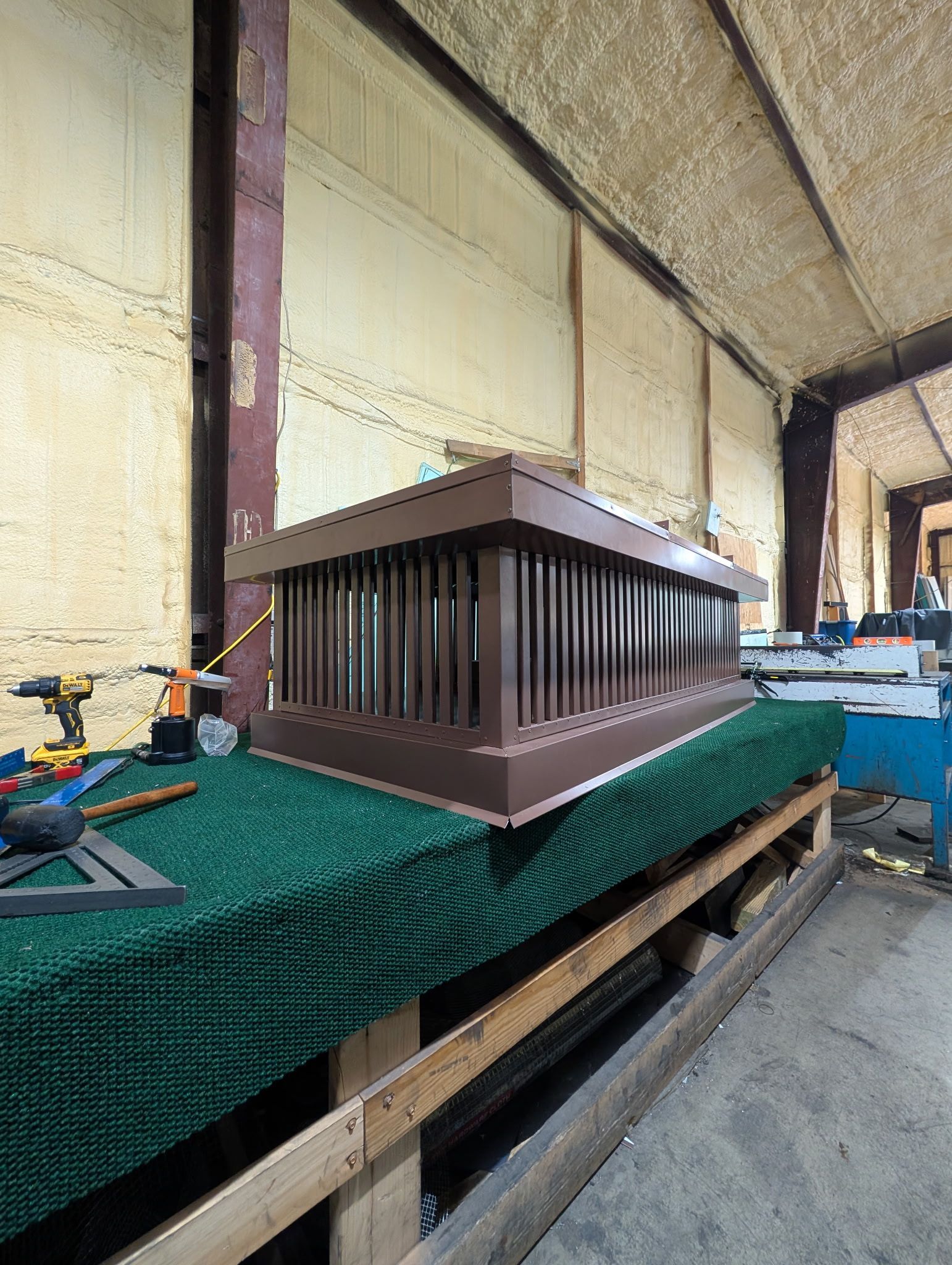 Small brown architectural model with columns on a green-covered workbench in a workshop or construction area