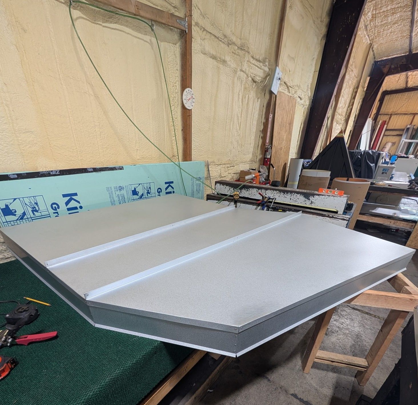 Large silver metal panel on a workshop table, with tools and insulation boards in a cluttered workshop.